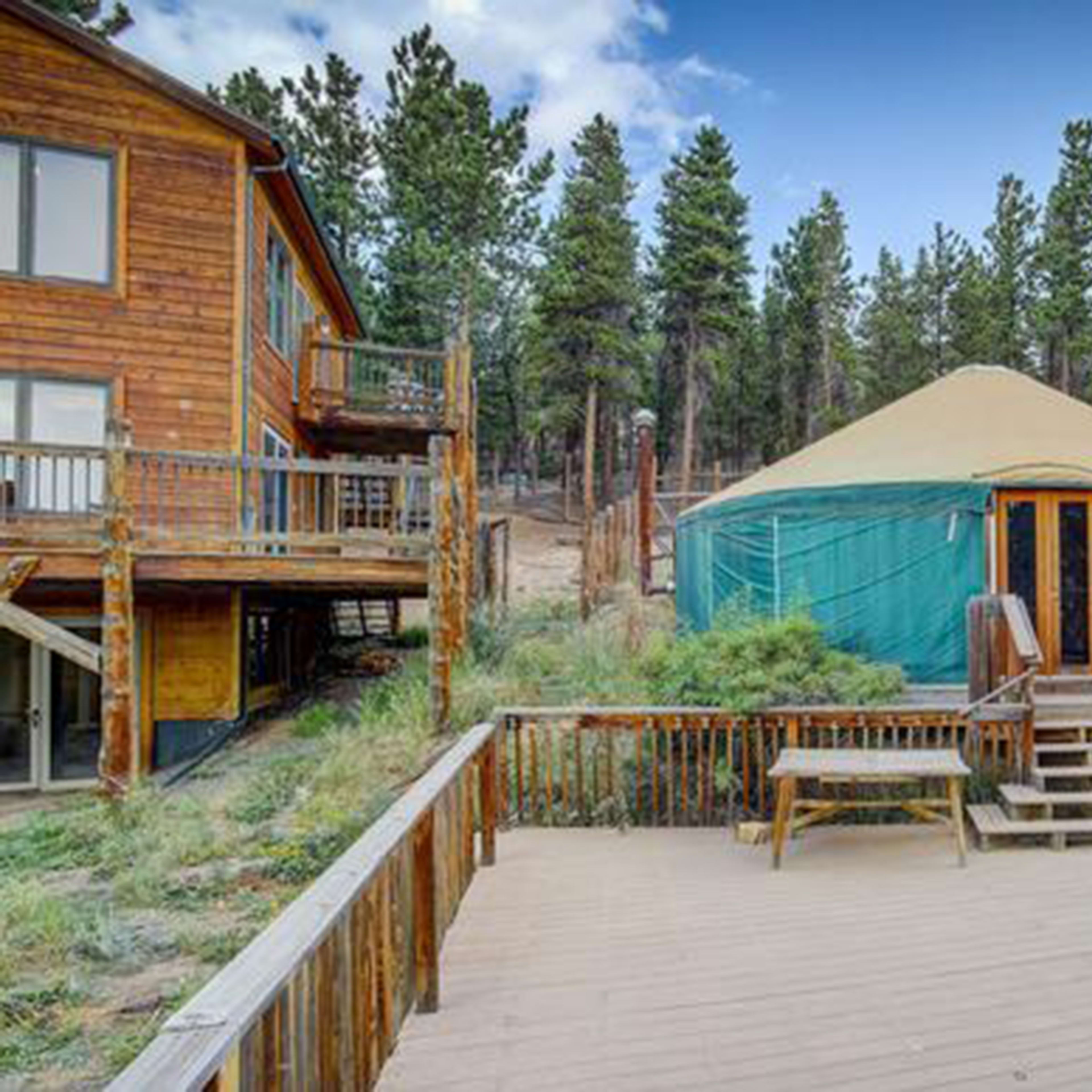A wooden deck leads to a green yurt surrounded by tall trees and a cabin in the background.