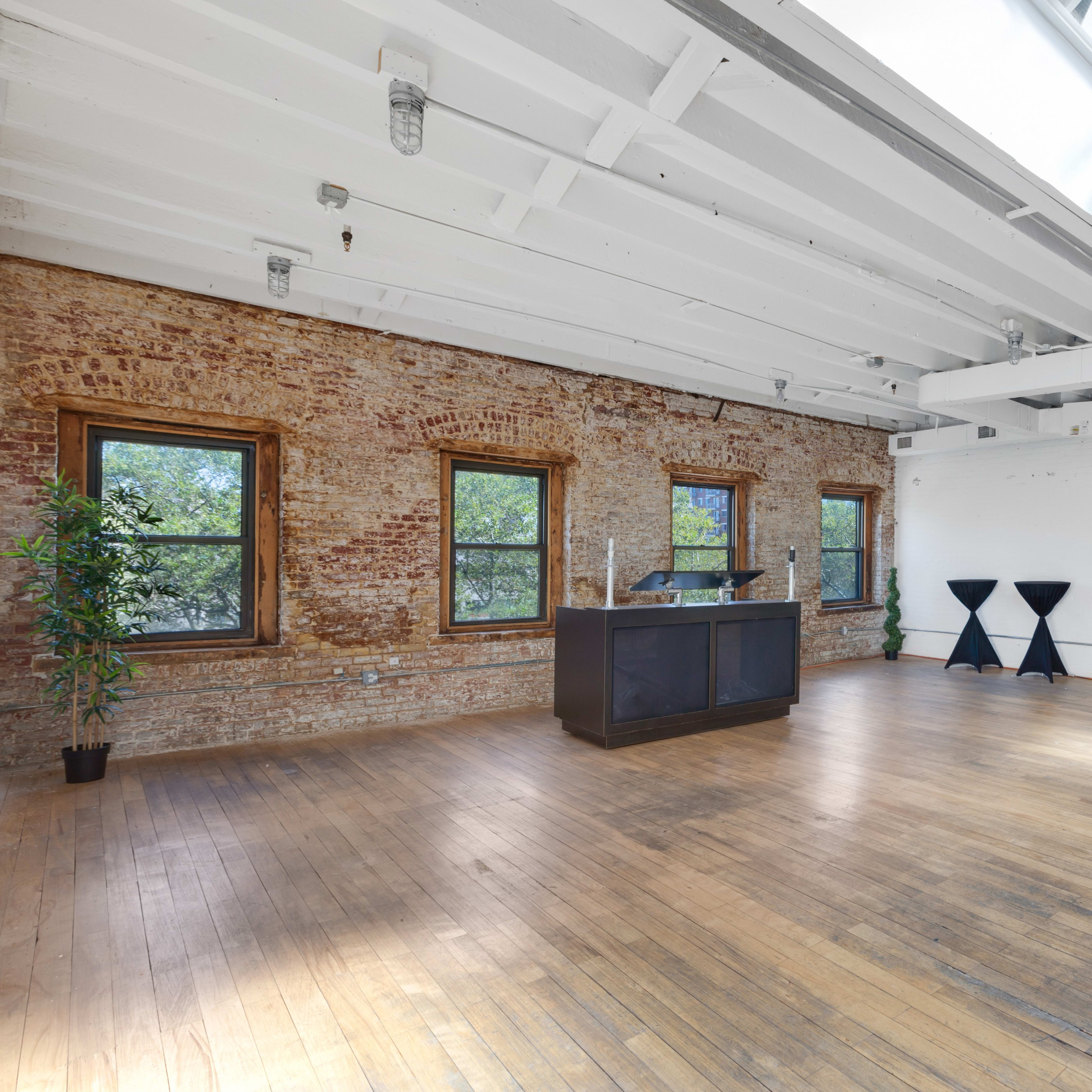A spacious, empty room with exposed brick walls, large windows, and wooden flooring, featuring a reception desk and two black display stands.