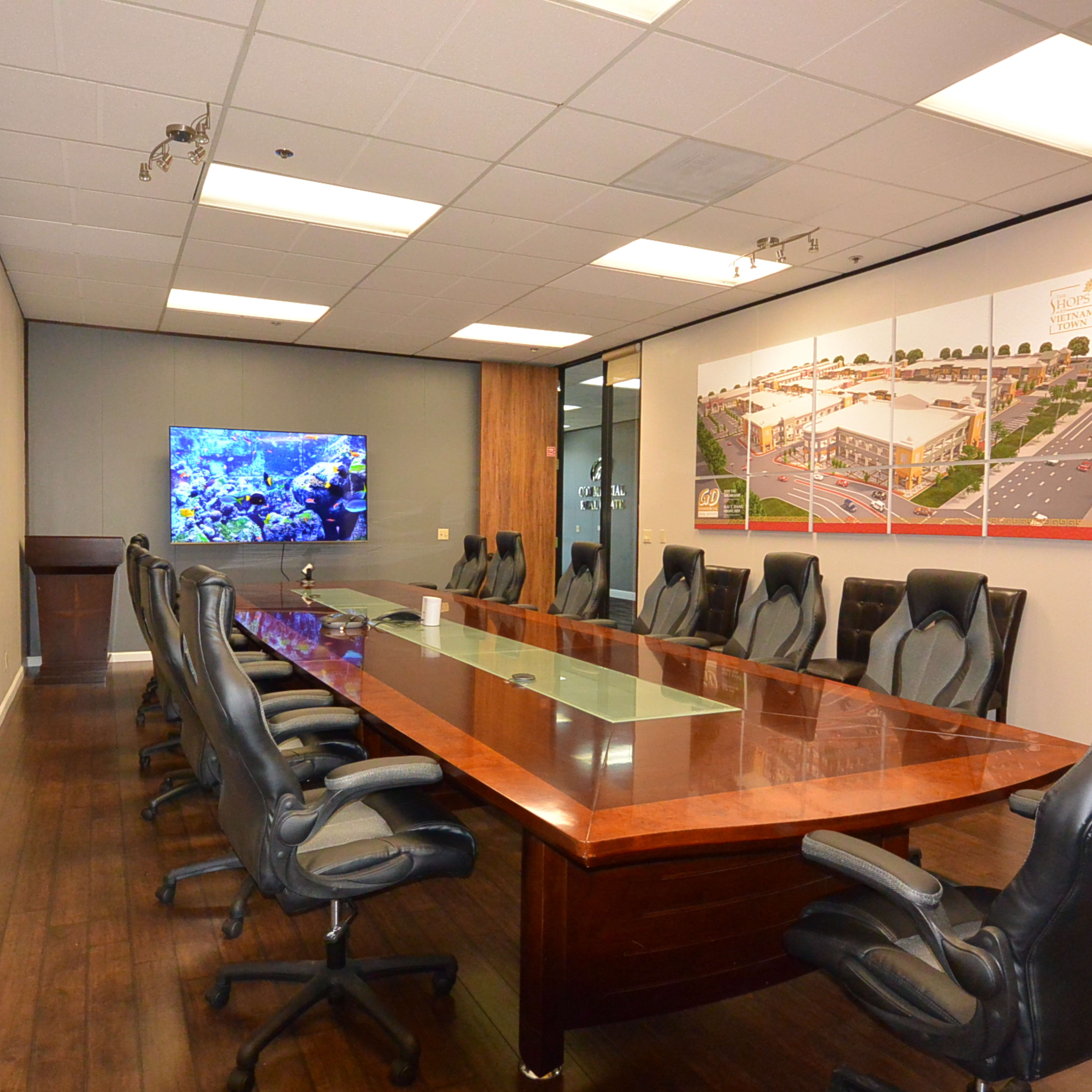 A large conference room features a long wooden table surrounded by black leather chairs, with a television and informational displays on the walls.
