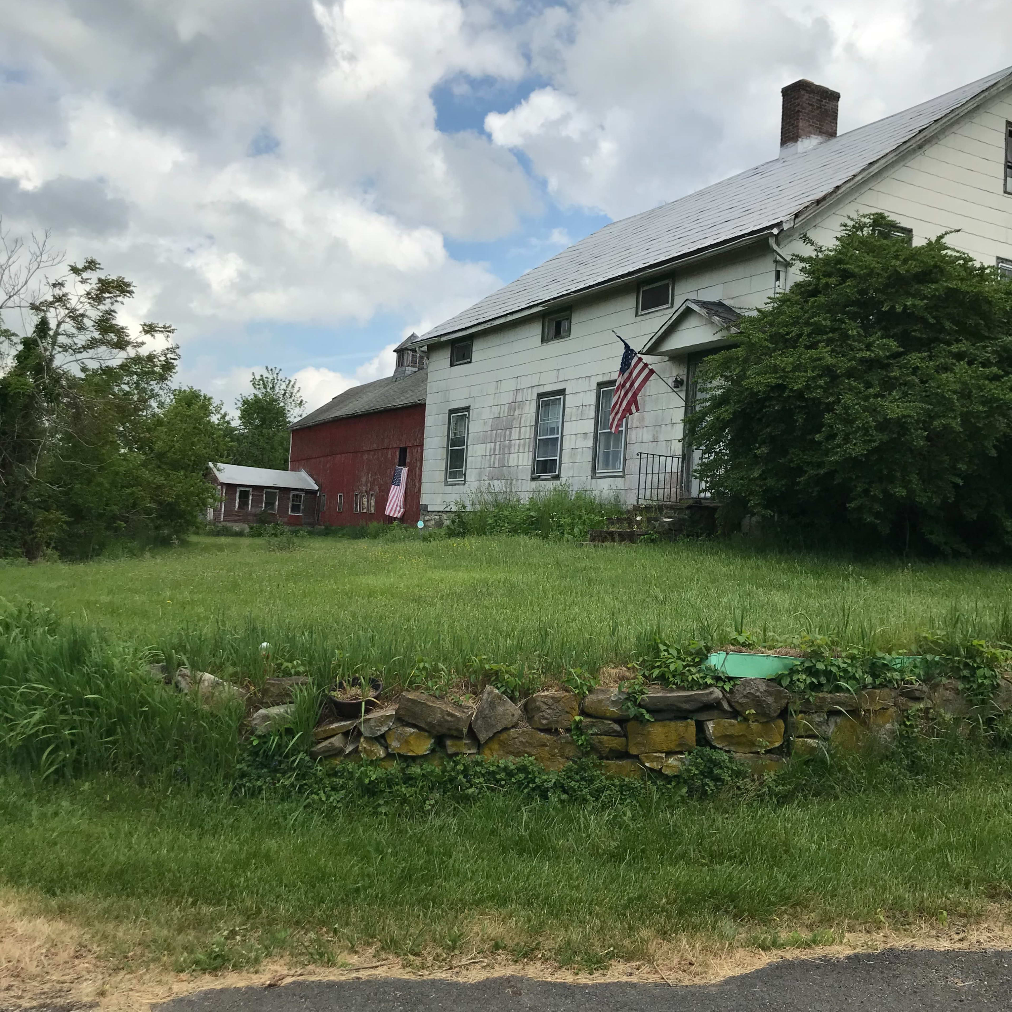 A white farmhouse with a gray roof and American flags hangs on the front, bordered by green grass and a stone wall, next to a red barn in a rural setting.