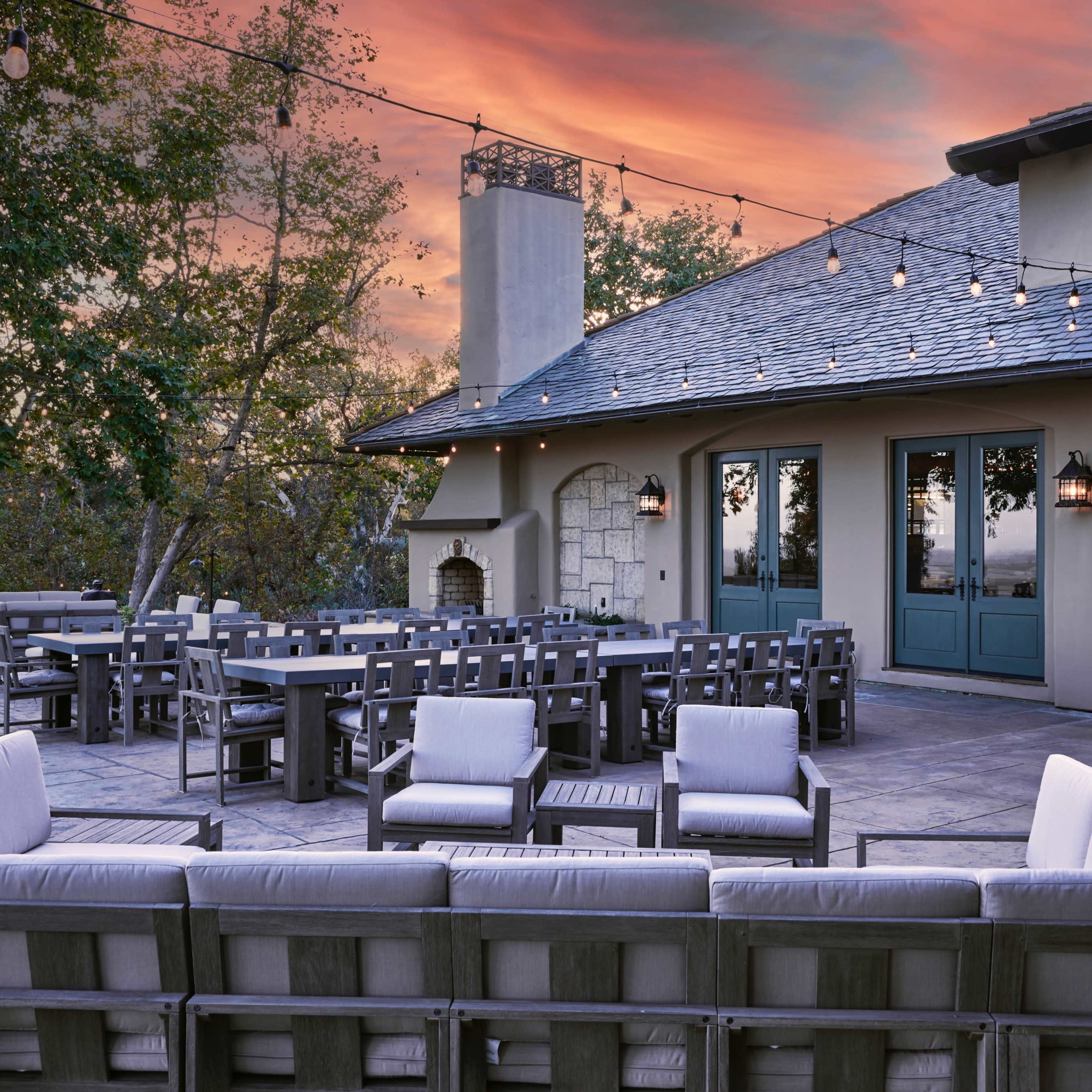 The scene features a patio with outdoor seating arranged in a circular layout, adjacent to a house with large windows and a stone fireplace, under a sky painted with sunset colors.