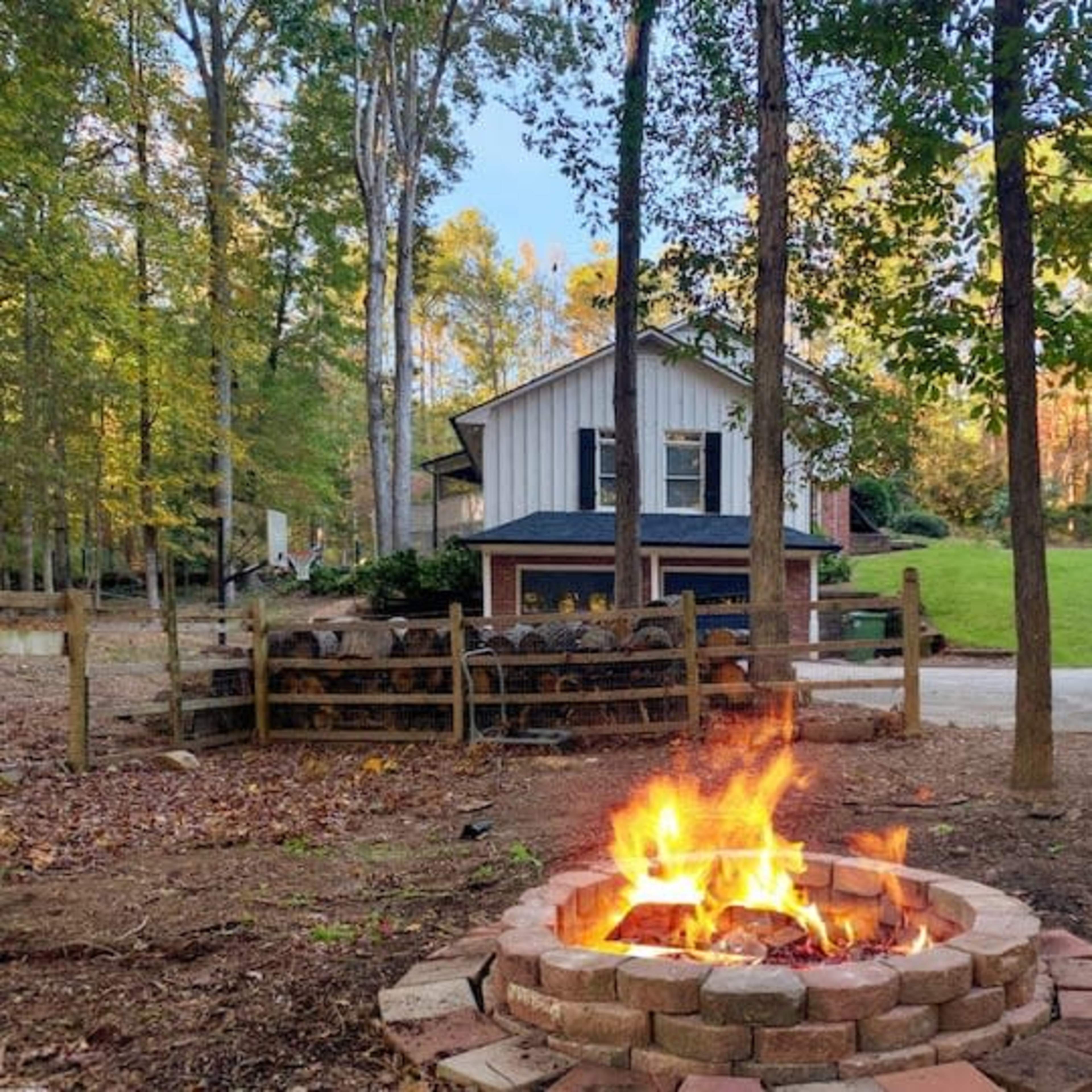 A stone fire pit with a vibrant fire is surrounded by trees, facing a two-story house in a wooded area.