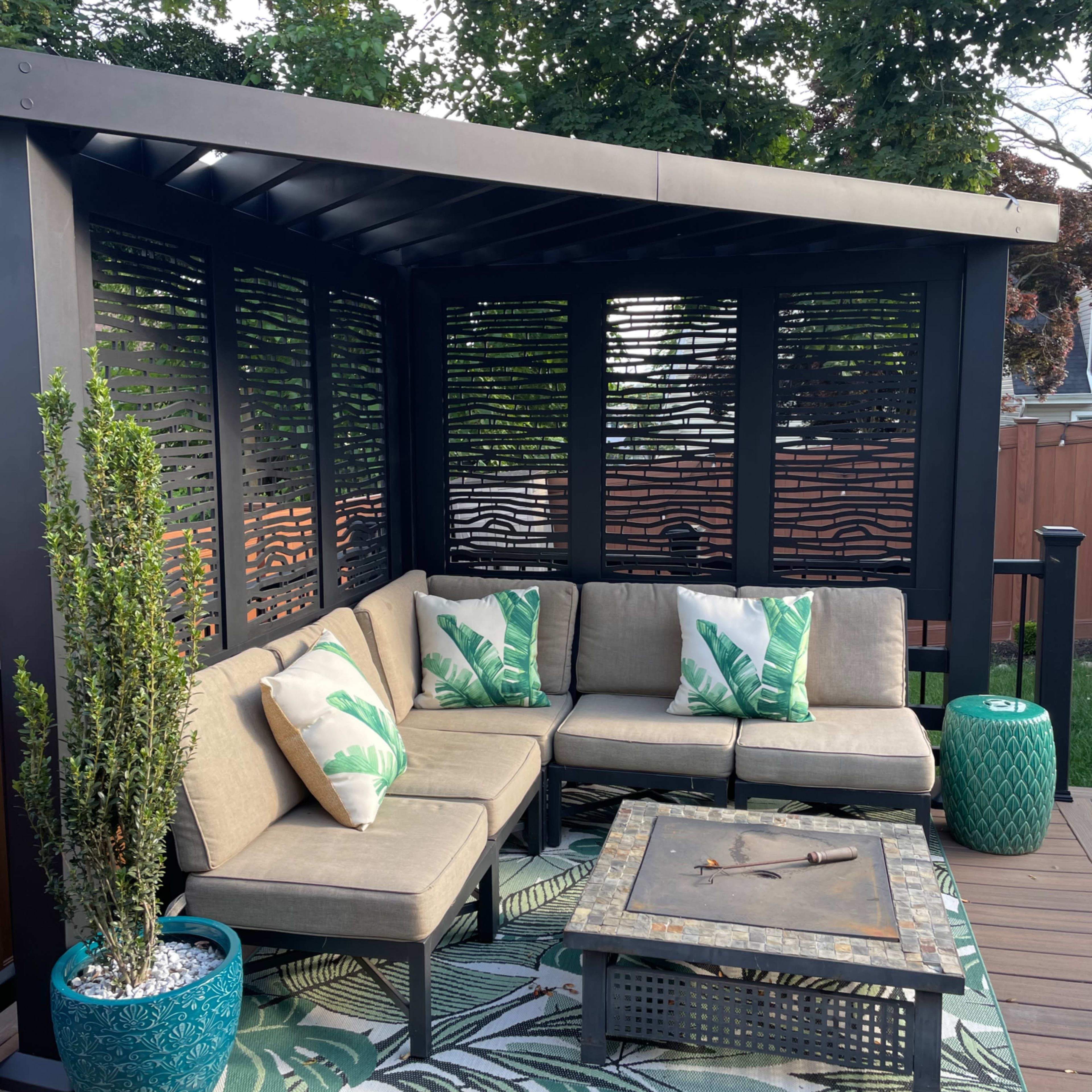 A covered outdoor seating area featuring a sectional sofa with green-accented pillows, a coffee table, and decorative plants on a patterned rug.