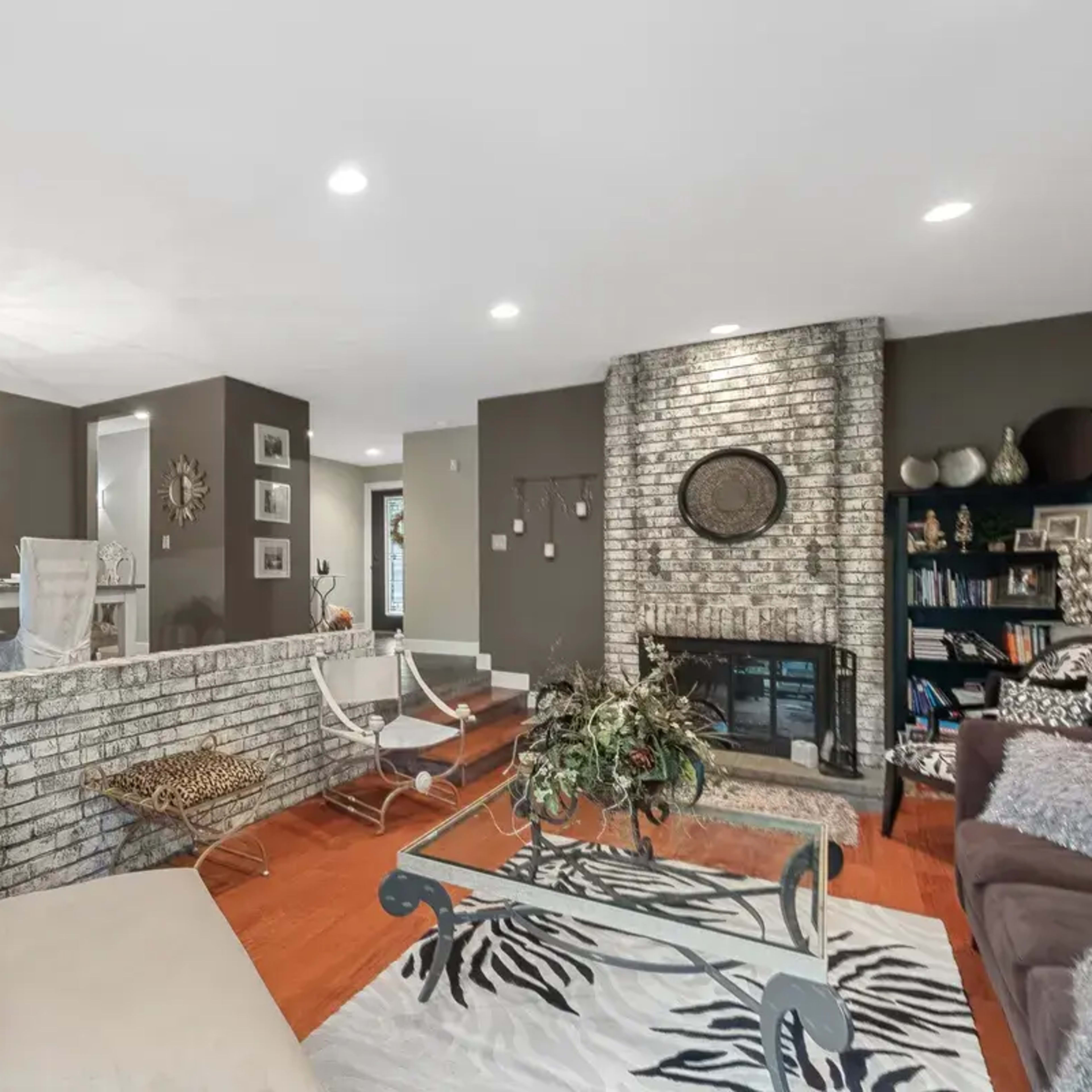 A living room with a stone fireplace, hardwood flooring, and a modern dining area visible in the background.