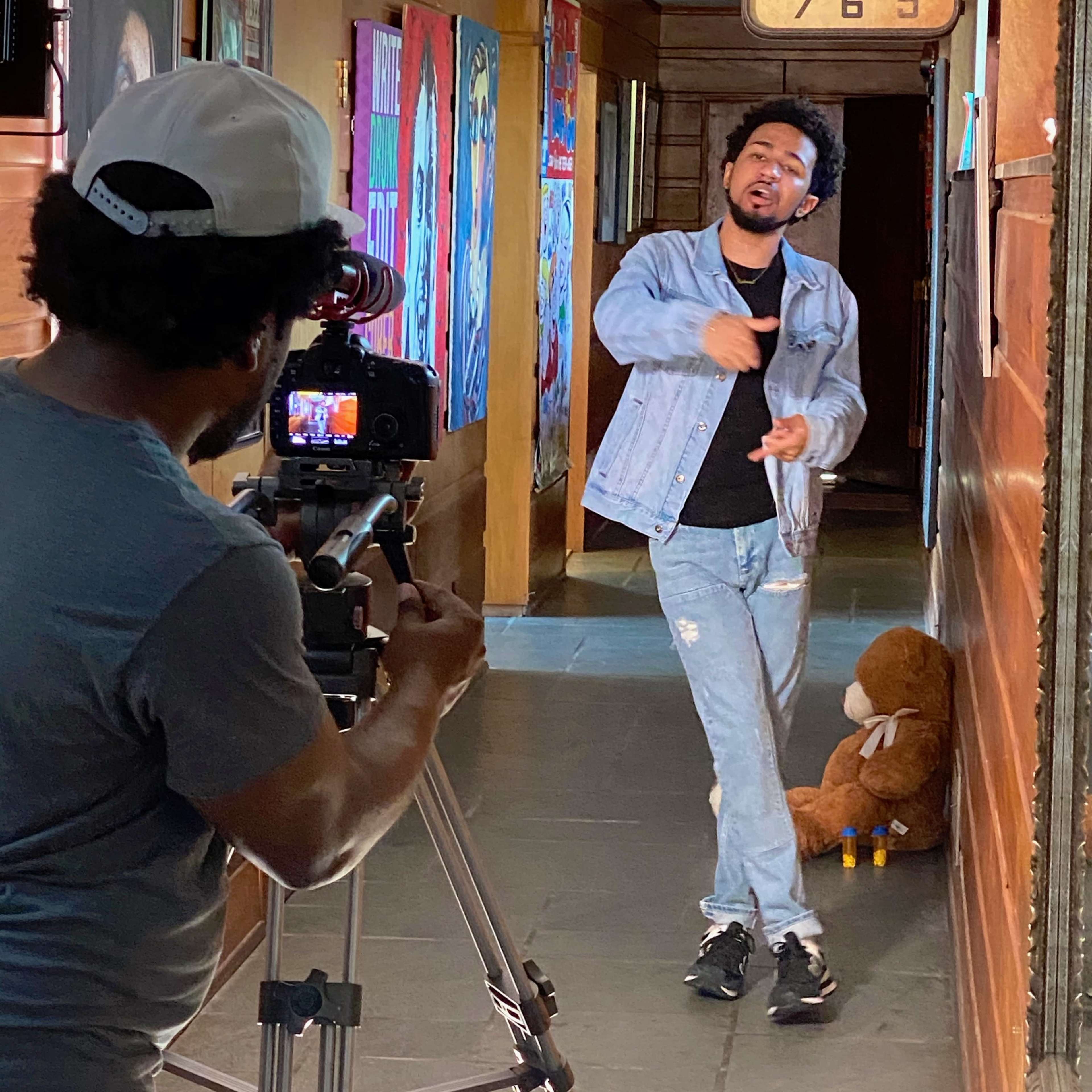 A person wearing a denim jacket performs in a hallway while another individual records the scene with a camera set on a tripod.