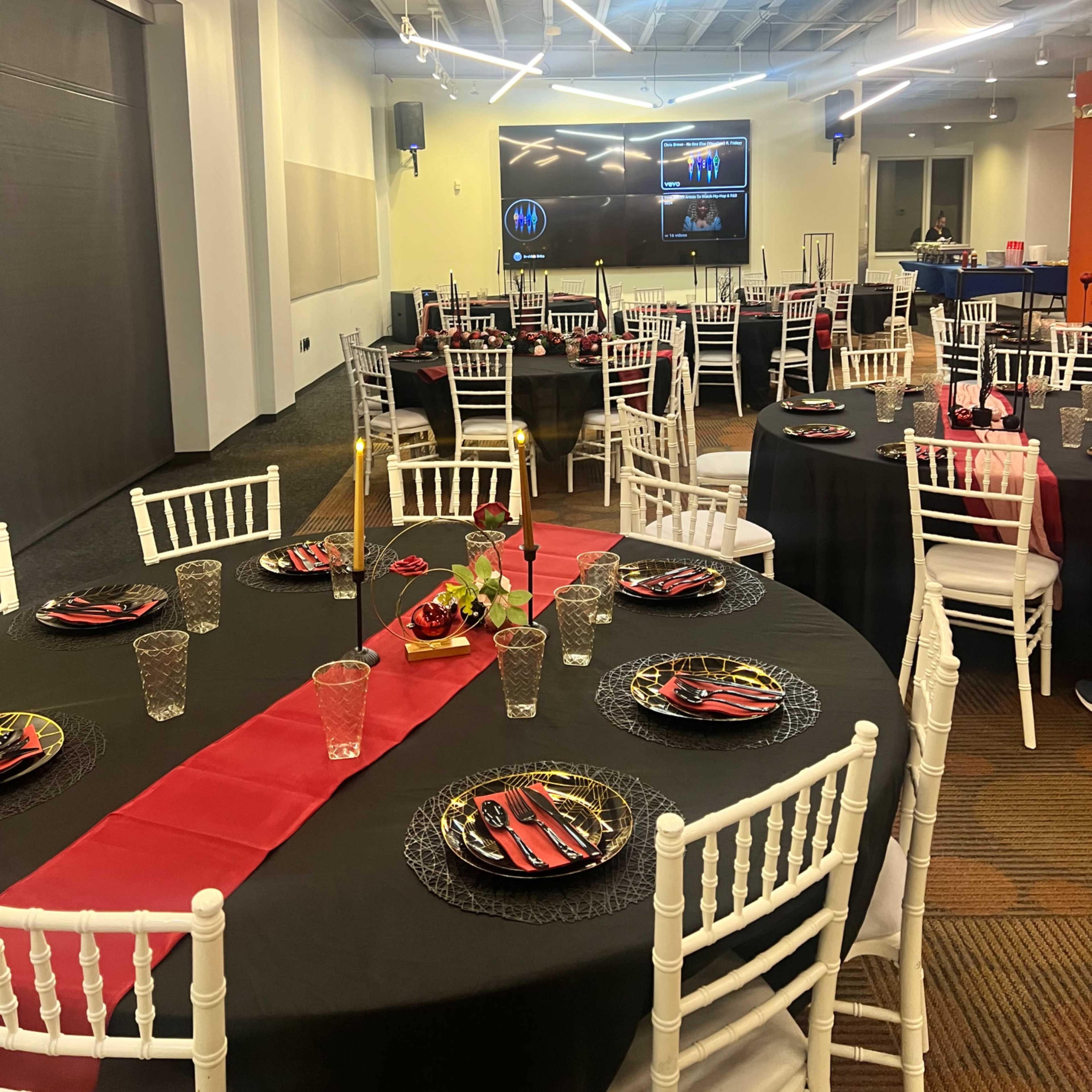 A banquet hall is set up with tables covered in black and red tablecloths, adorned with plates and glasses, and chairs placed around them, while a screen displays images at the front of the room.