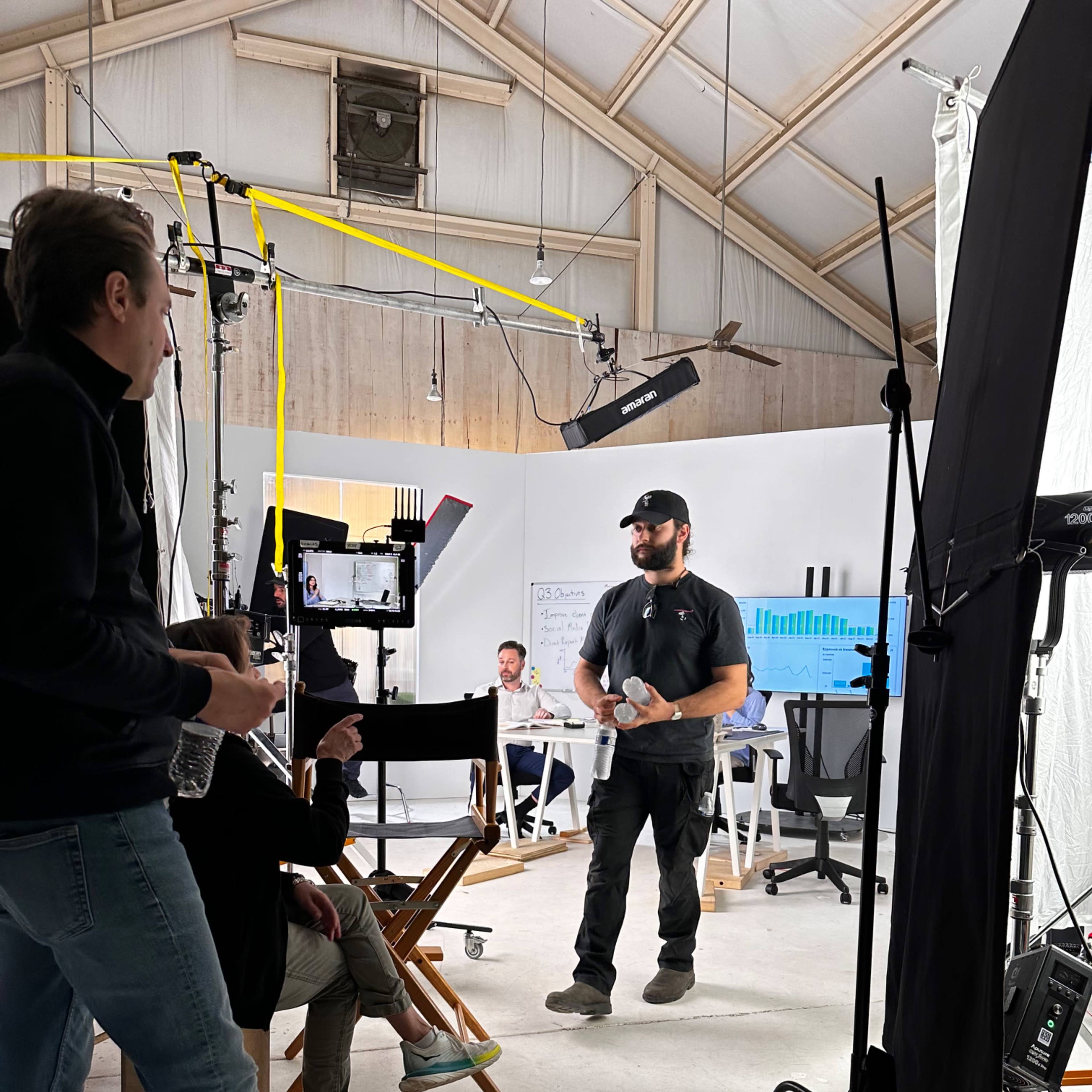 A production team is set up in a studio, with one person standing and presenting to a small audience seated nearby, while various cameras and lighting equipment are arranged around the space.