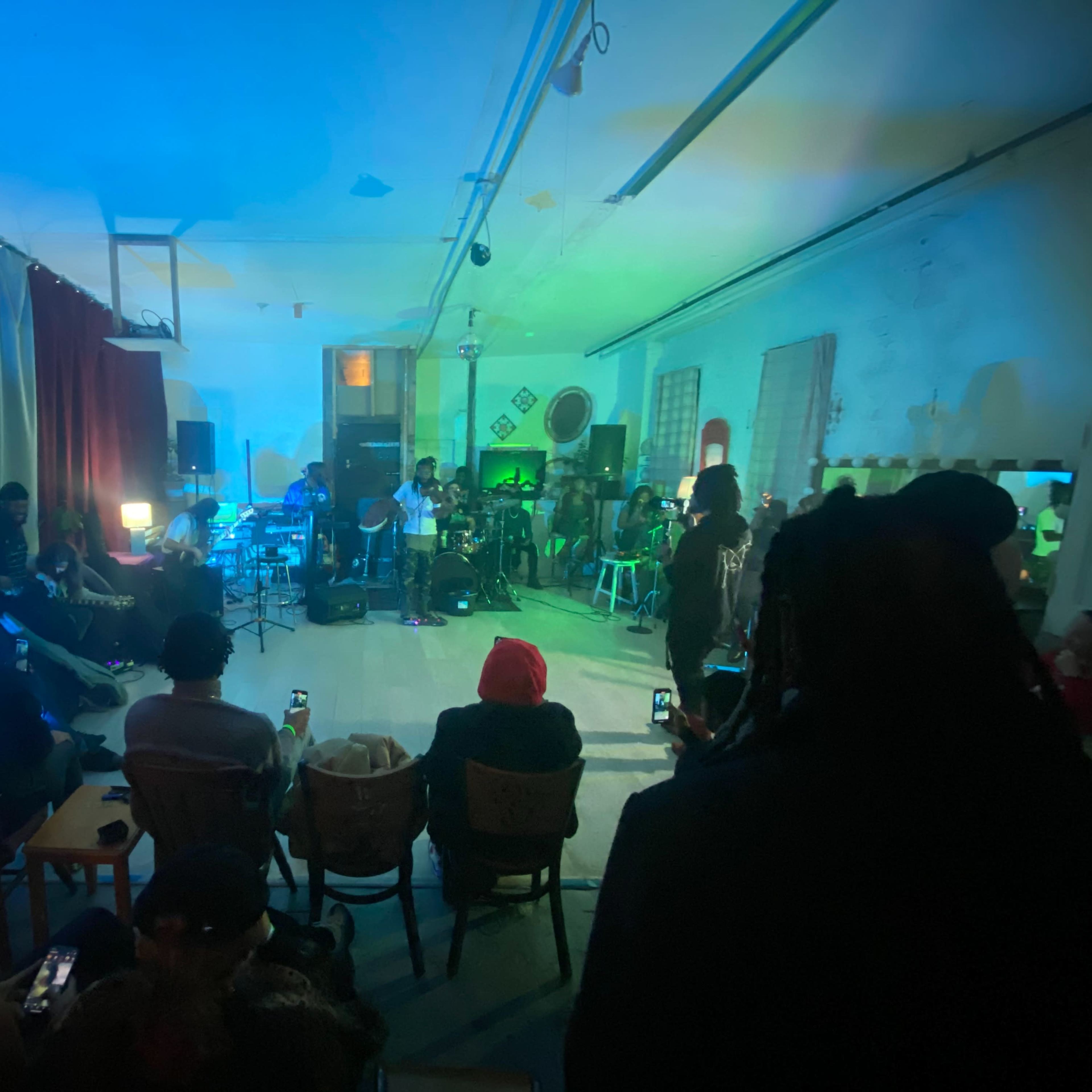 A small, dimly lit performance space is filled with an audience seated in chairs while a live band plays on stage under blue and green lighting.
