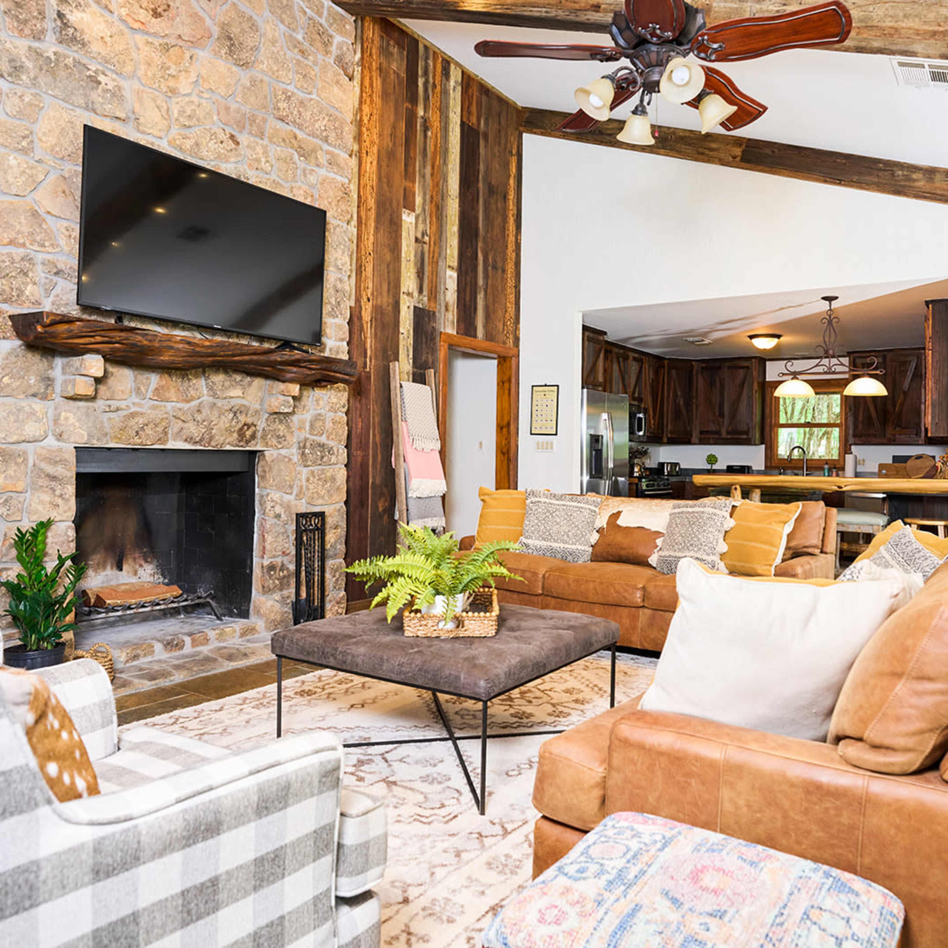 The living room features a stone fireplace, a large flat-screen TV mounted above it, and a mix of seating options including a leather sofa and patterned chairs.