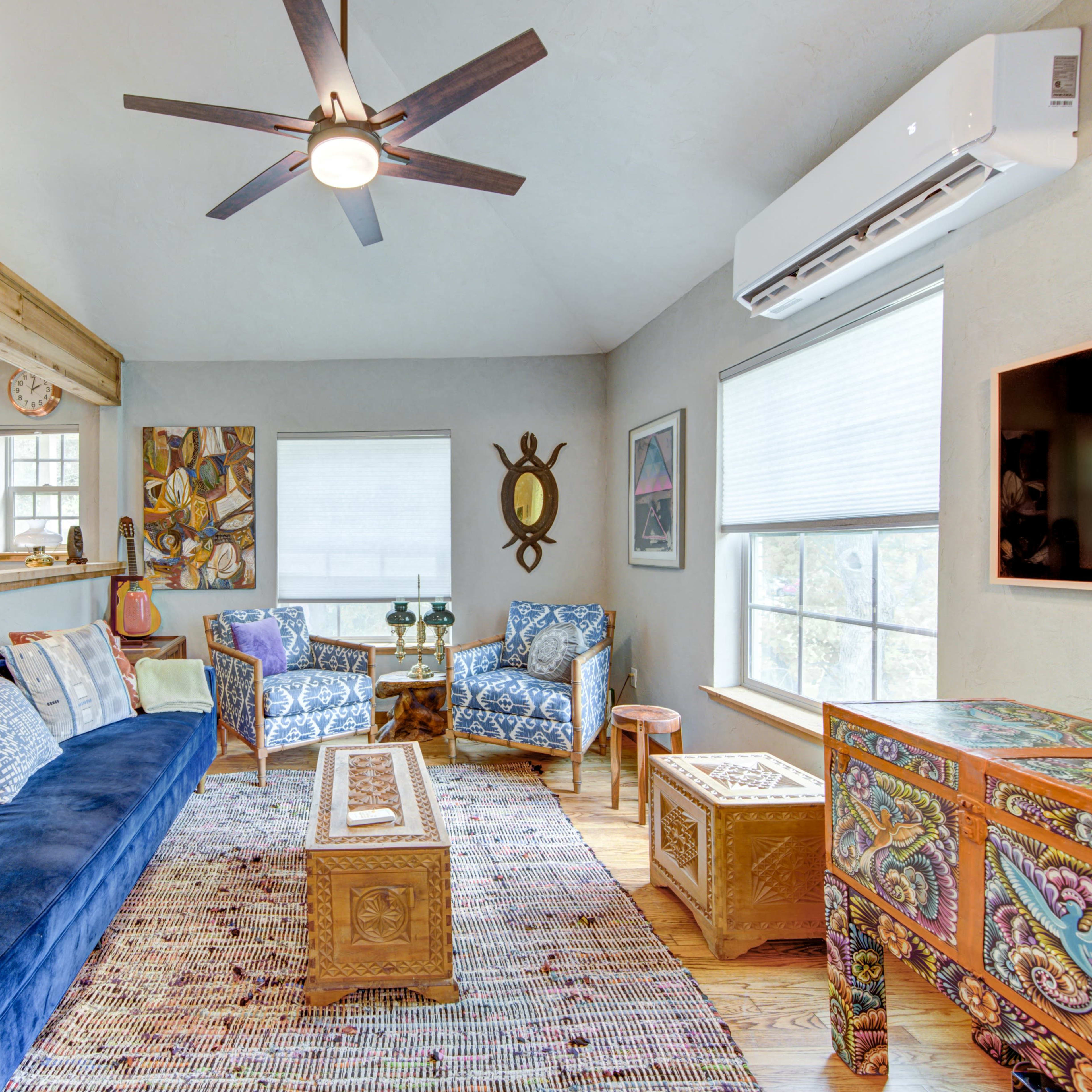 A cozy living room featuring a blue sofa, patterned armchairs, and decorative wooden furniture under a ceiling fan, with natural light streaming through the windows.