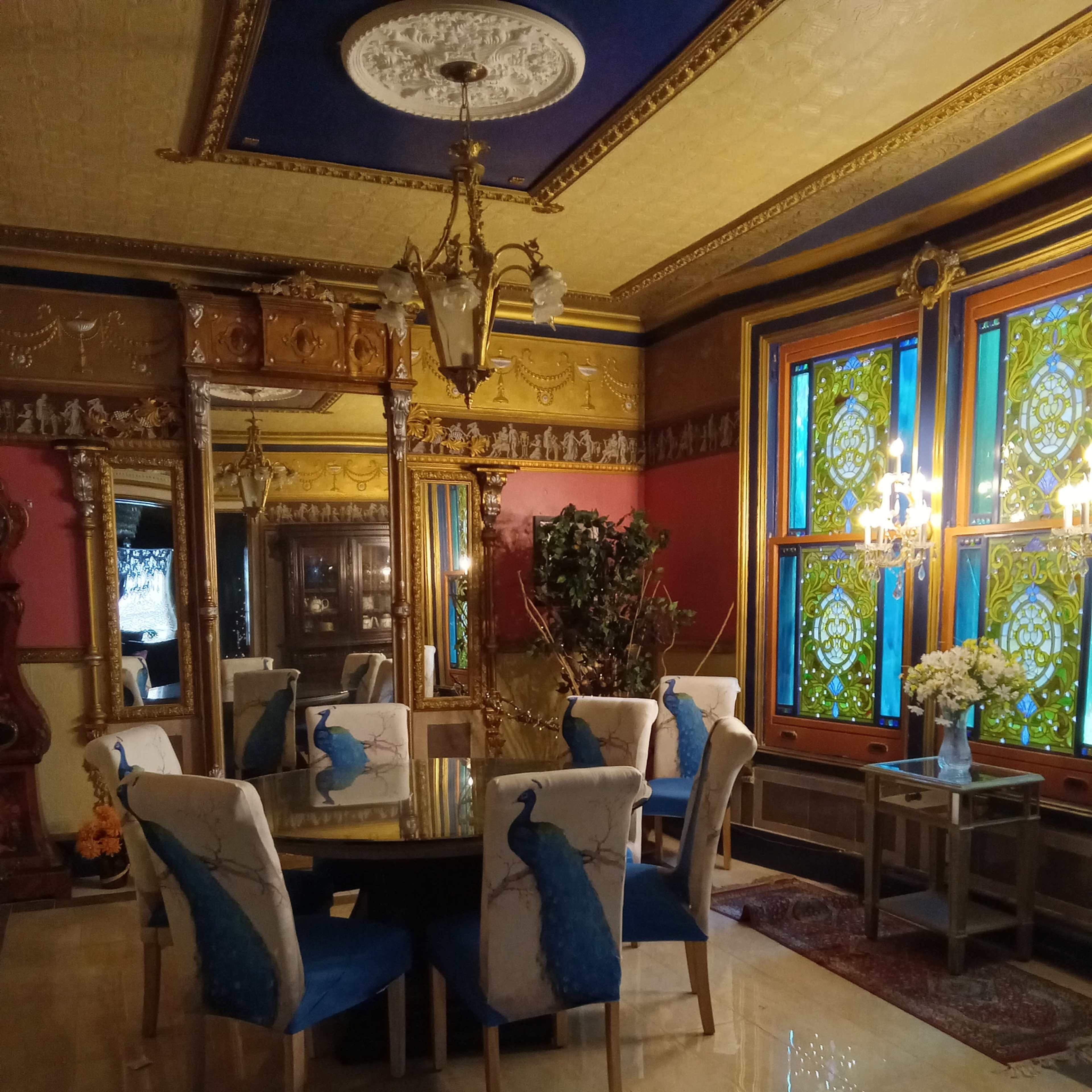 An elegantly decorated dining room featuring a round table surrounded by chairs with peacock designs, illuminated by a chandelier and adorned with colorful stained glass windows.