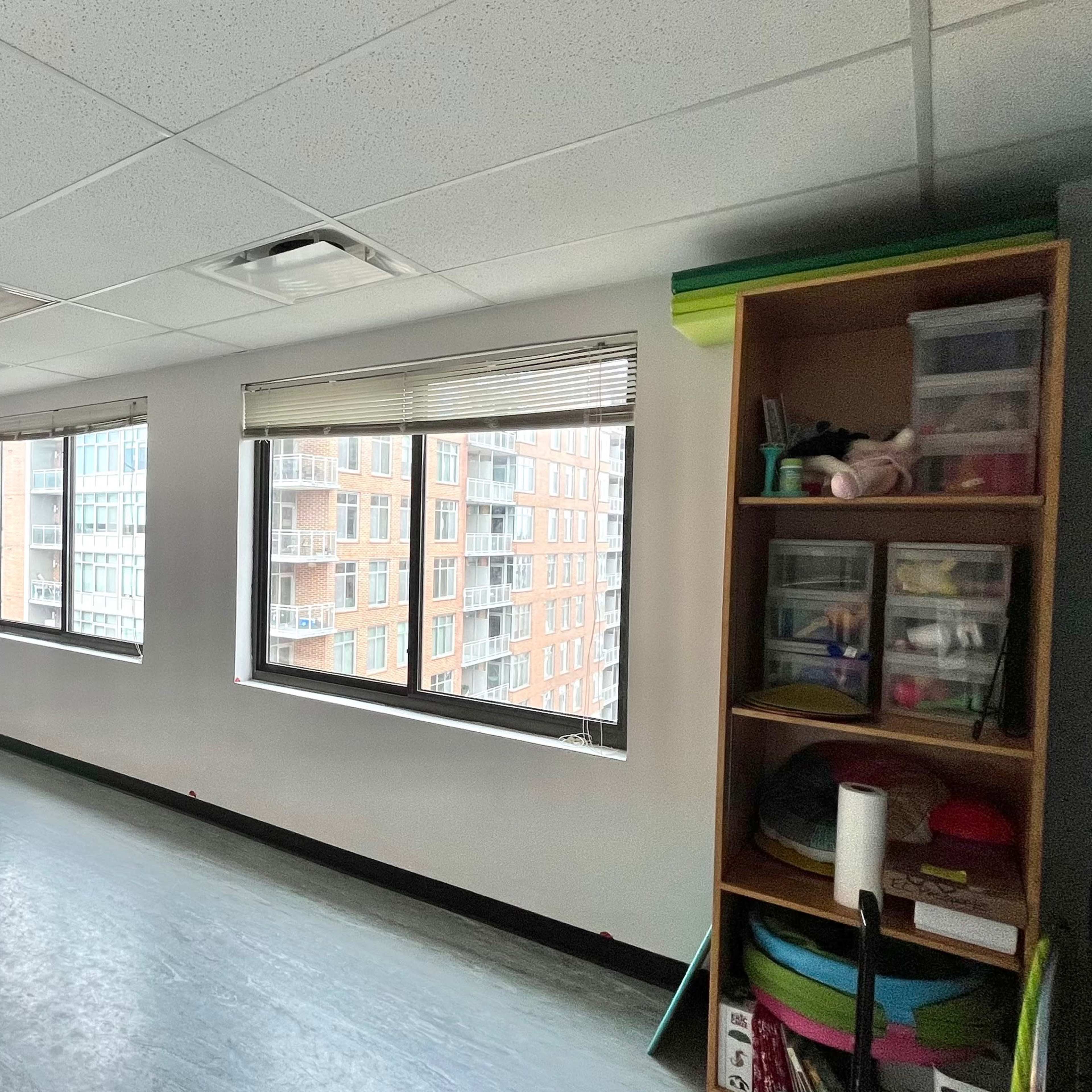 An empty dance studio with large windows, a mirrored wall, and a shelving unit containing various items.