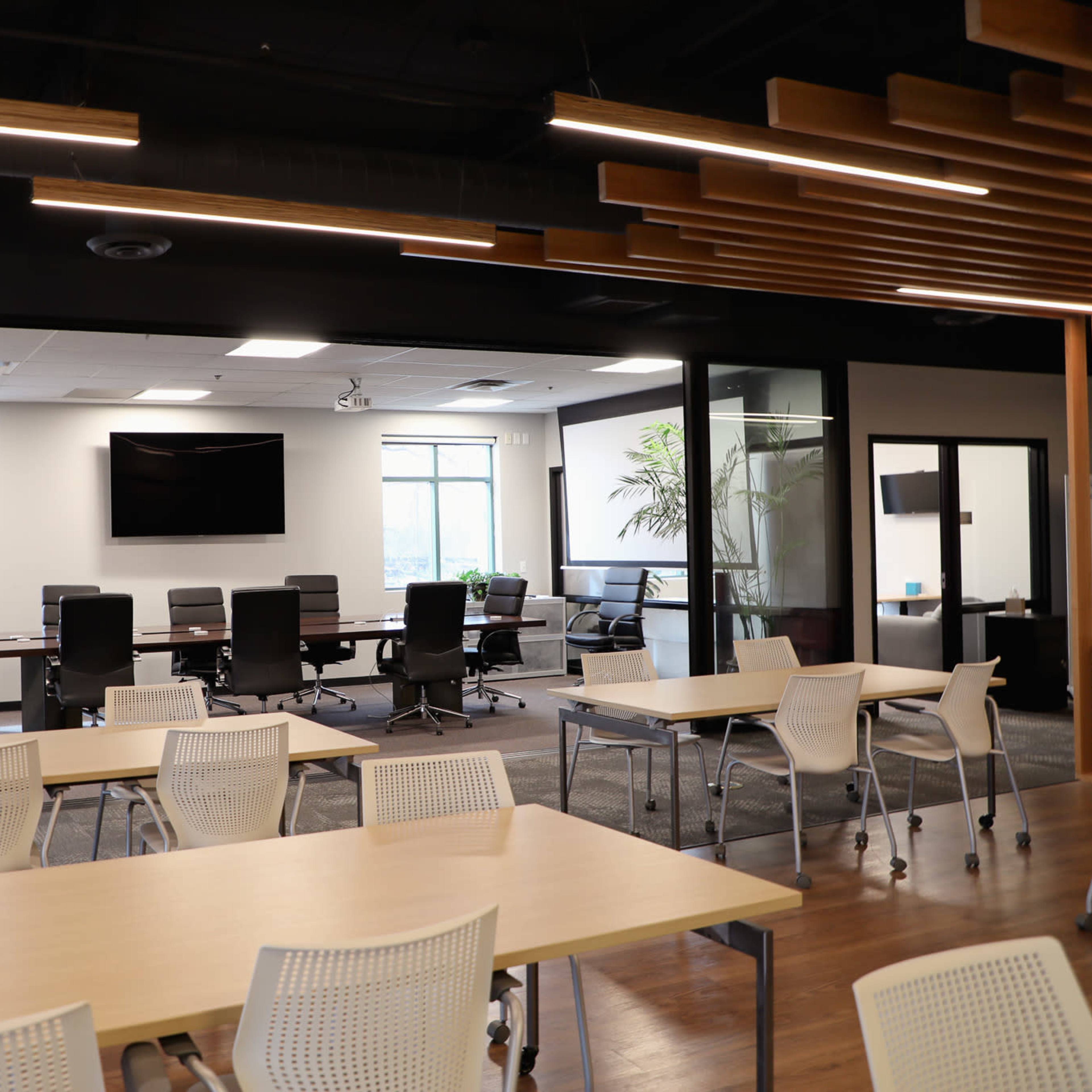 A modern office space features several tables and chairs in the foreground, with a glass-walled conference room visible in the background.