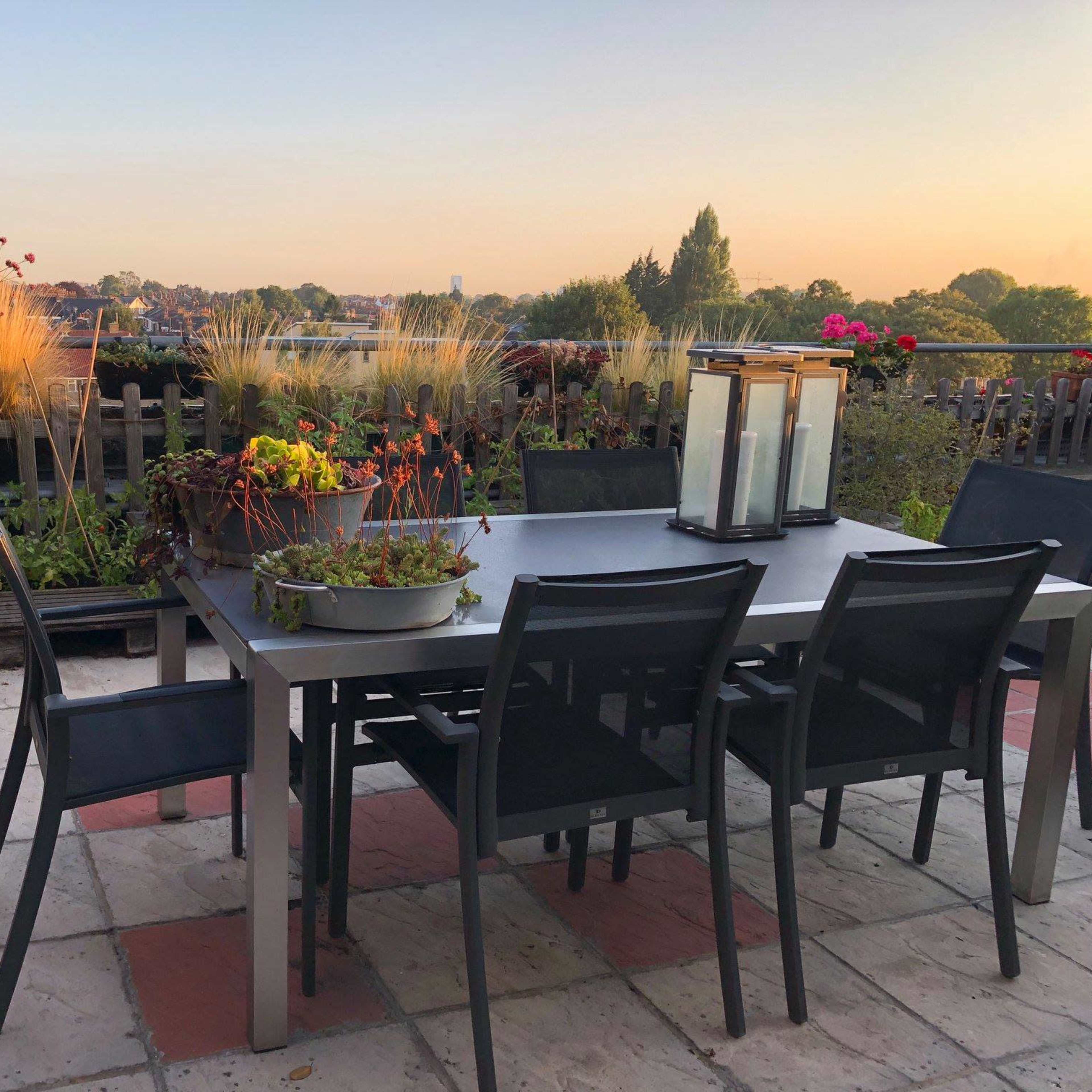 A terrace dining area with a table and chairs surrounded by various potted plants and a lantern, set against a backdrop of a sunset sky.