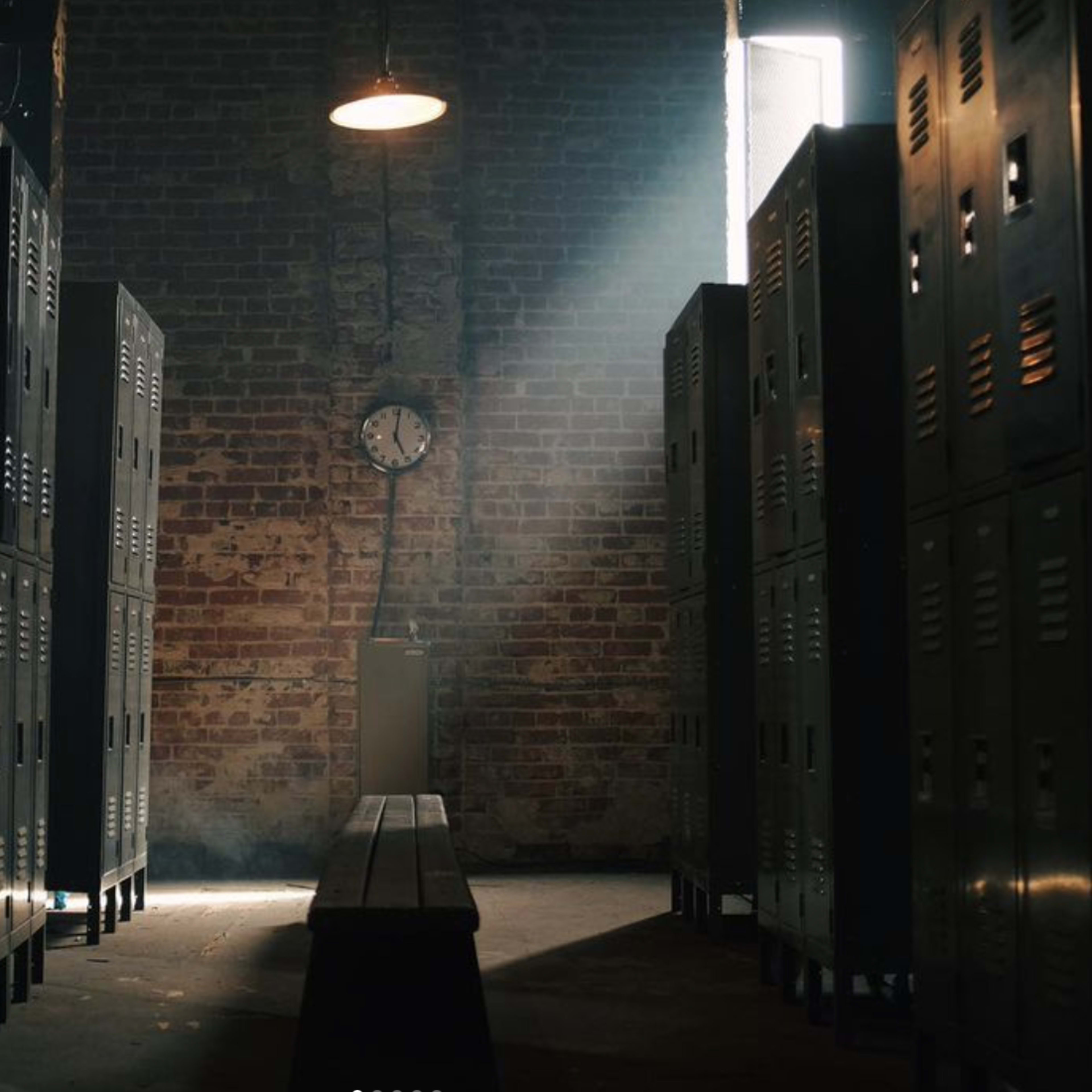 A dimly lit locker room with metal lockers lining the walls and a wooden bench in the center, illuminated by a beam of light from a window.