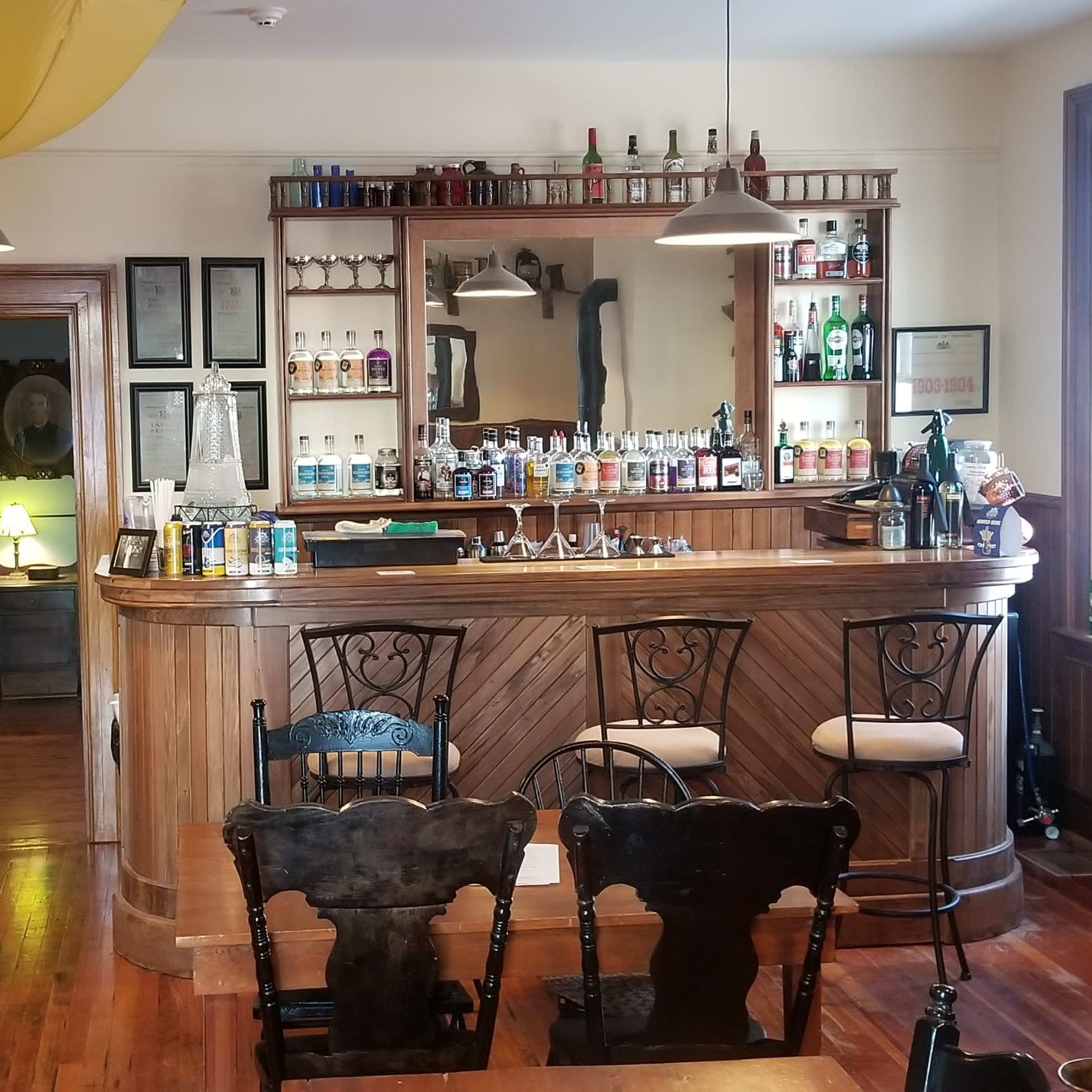 A well-furnished bar area with a wooden counter, several rows of bottles on shelves, and wooden tables and chairs arranged around the space.