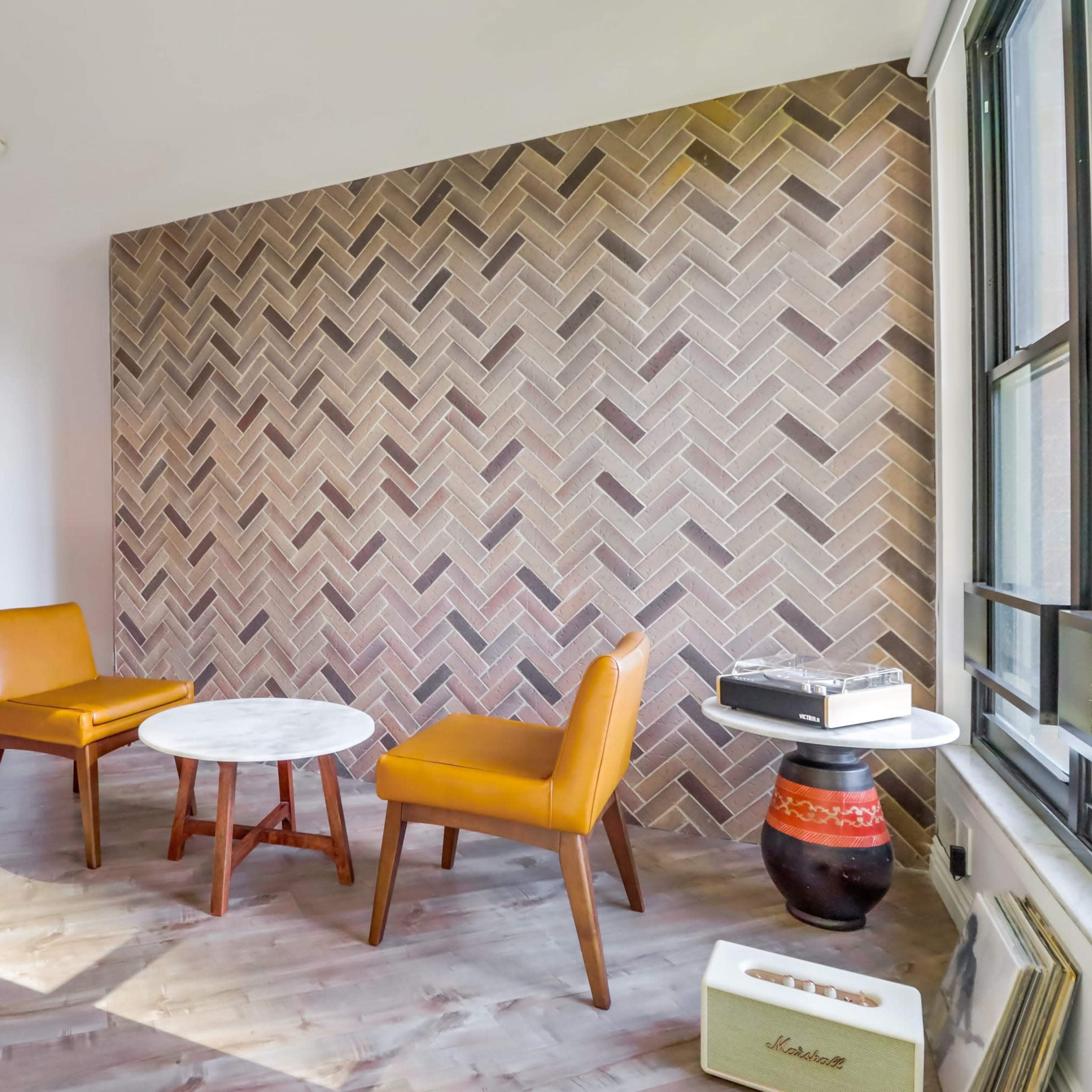 A modern room with a herringbone-patterned accent wall, two yellow chairs, a round marble table, and a small lamp.