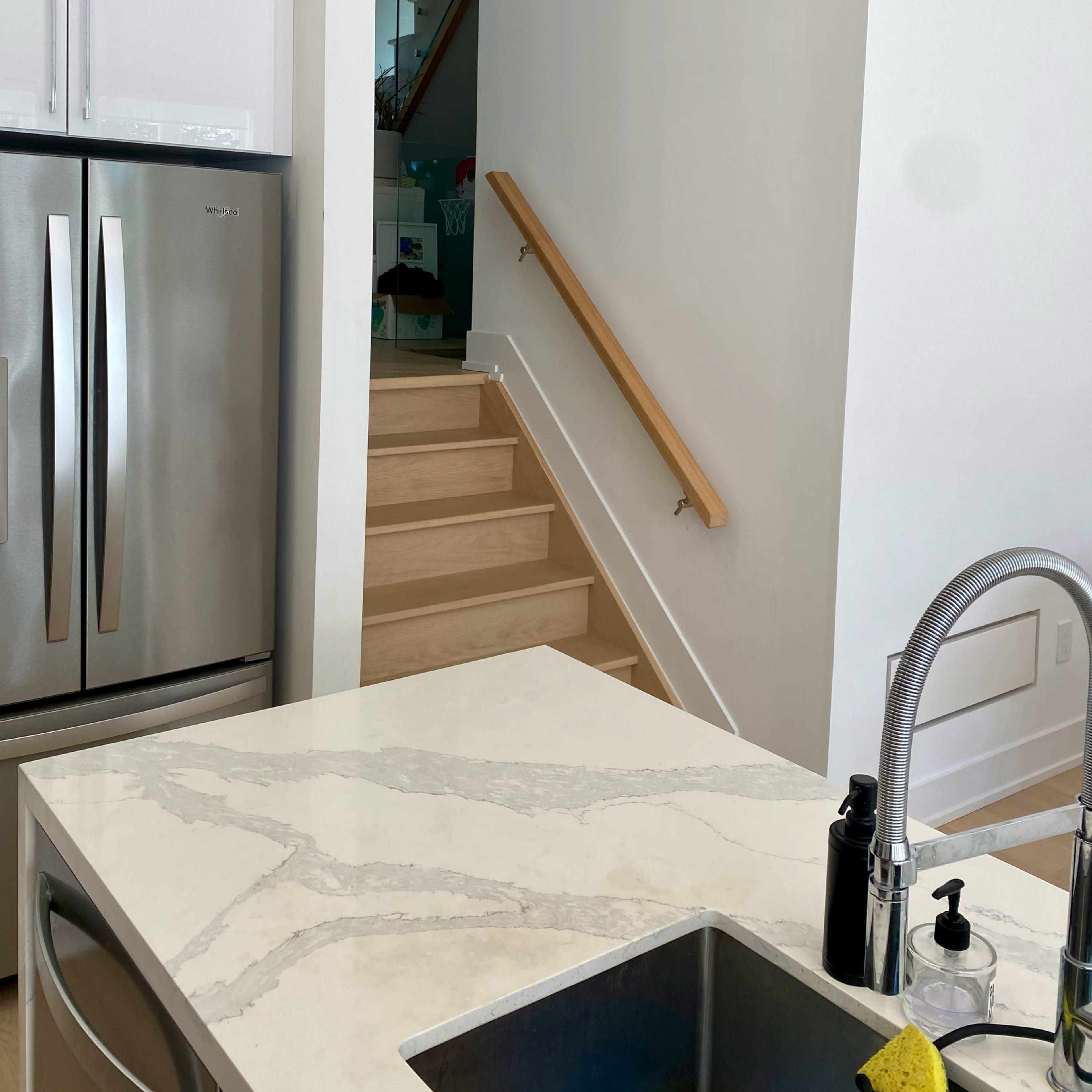 A modern kitchen featuring a stainless steel refrigerator and a marble countertop, with a staircase visible in the background.
