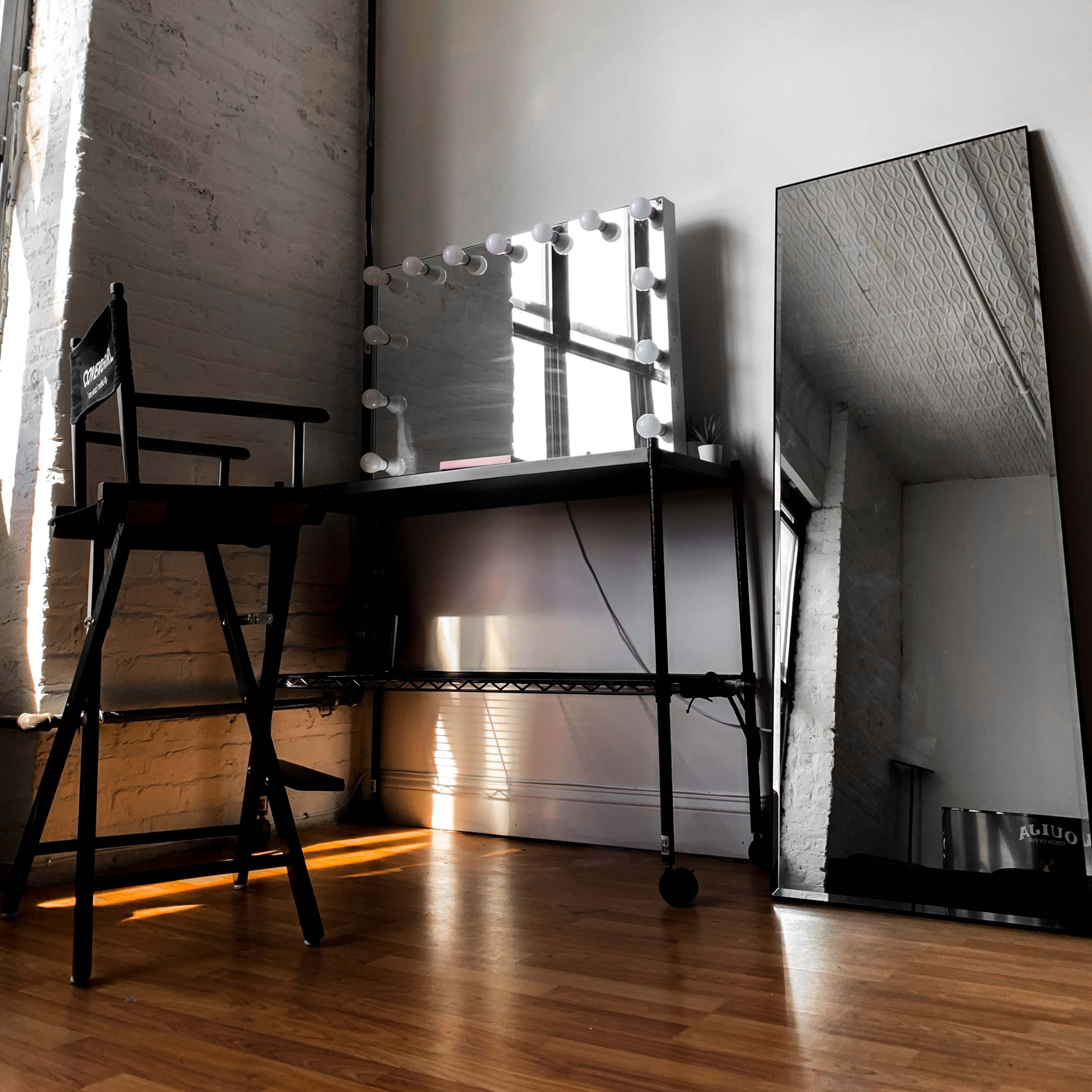A bright room with a wooden floor, featuring a makeup vanity with lights and a director's chair next to a tall mirror against the wall.