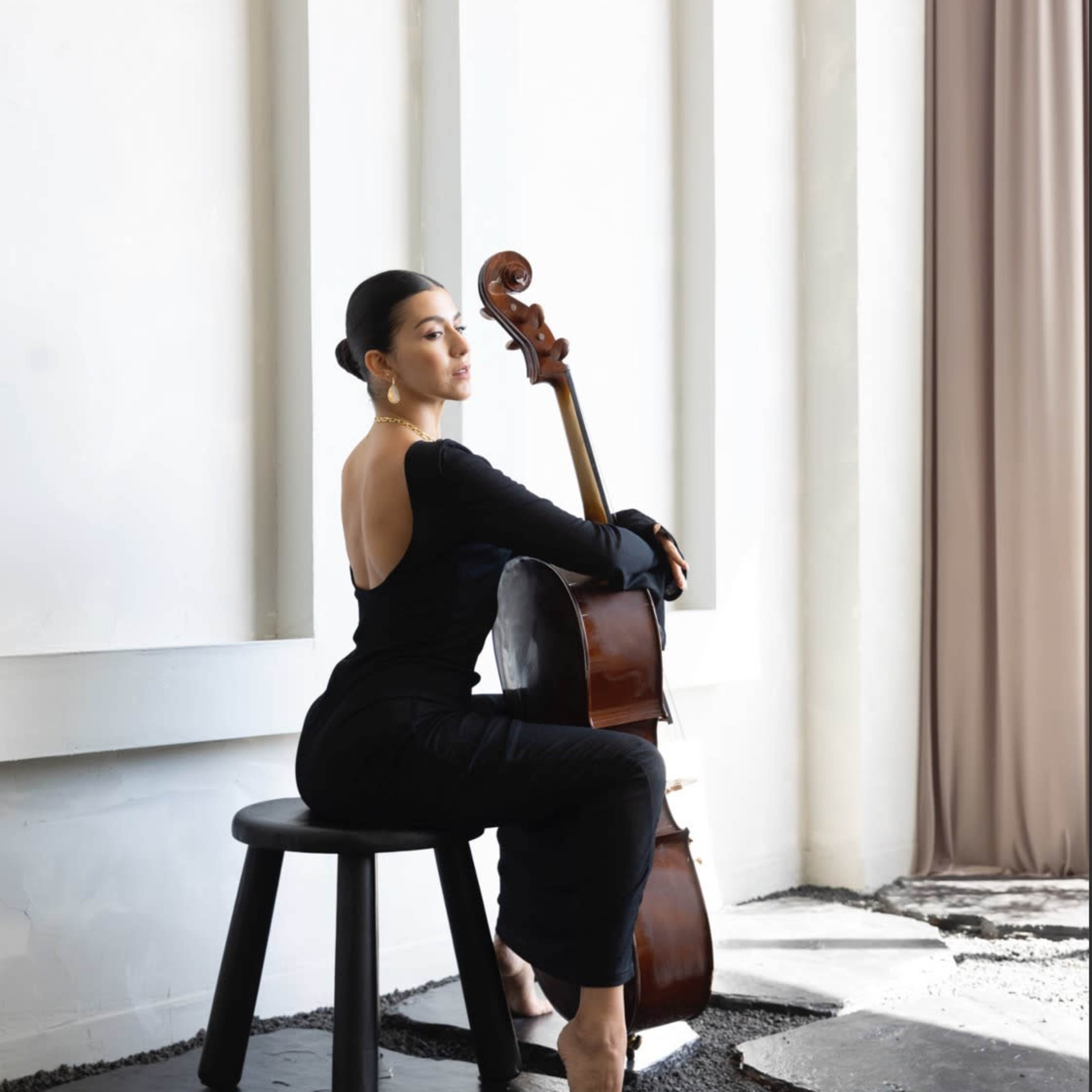 A woman dressed in black sits on a stool, elegantly posed with a cello beside her in a minimalist interior setting.
