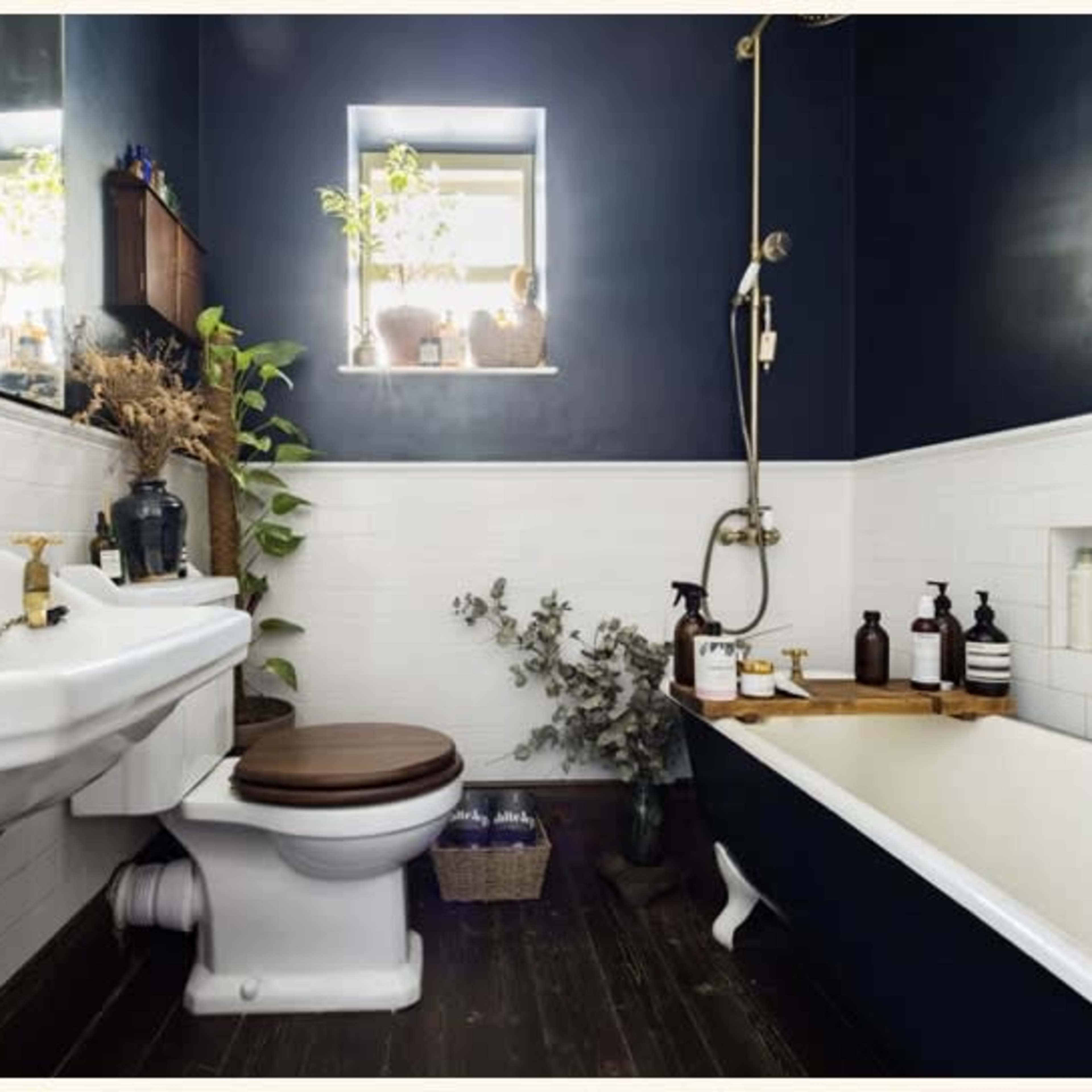A compact bathroom featuring white tiled walls, a wooden floor, a porcelain toilet, a vintage-style sink, a clawfoot bathtub, and shelves with plants and toiletries.