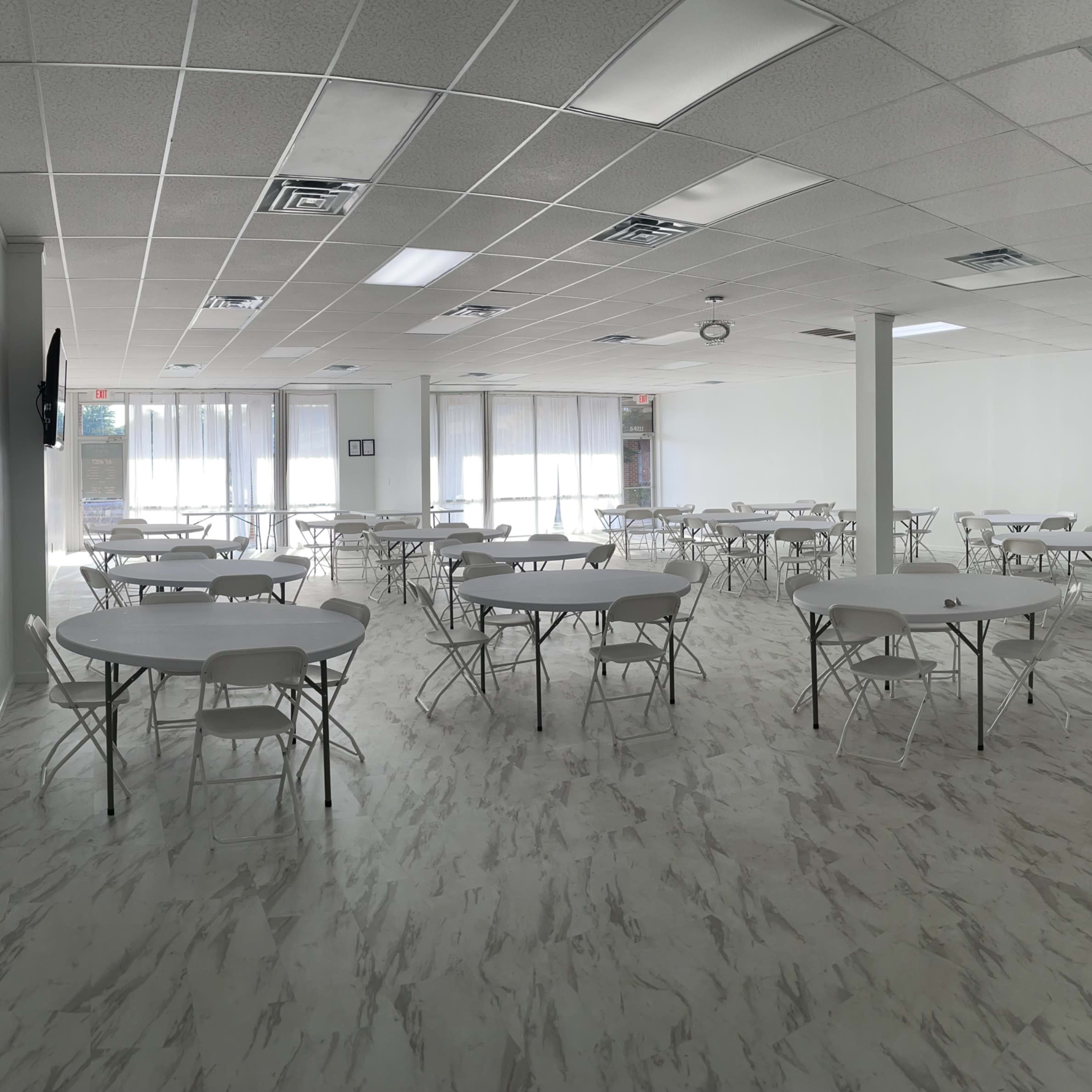 A spacious room with white walls and a patterned floor, featuring multiple round tables surrounded by chairs.