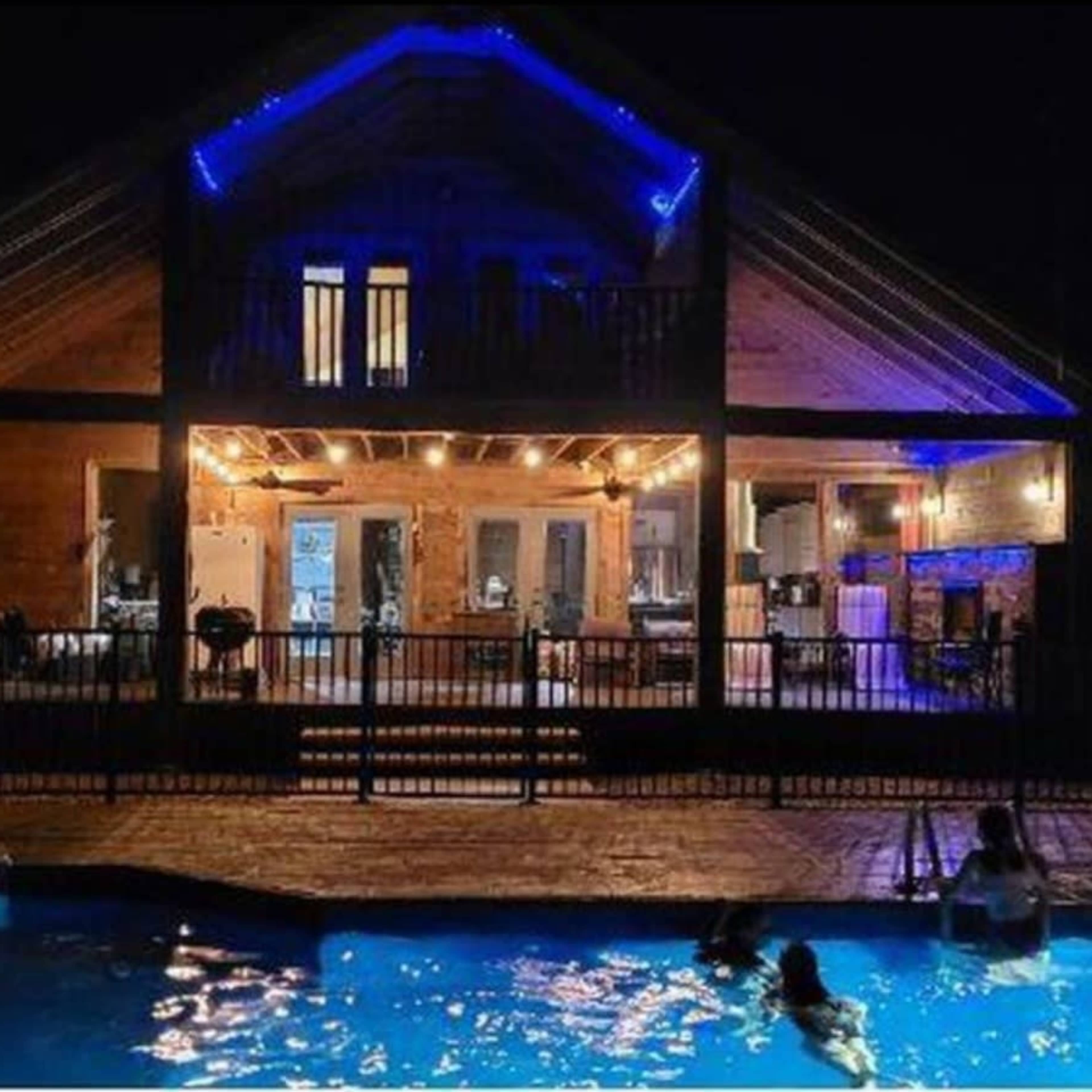 A large wooden house with illuminated features stands behind a swimming pool at night, with people swimming in the water.
