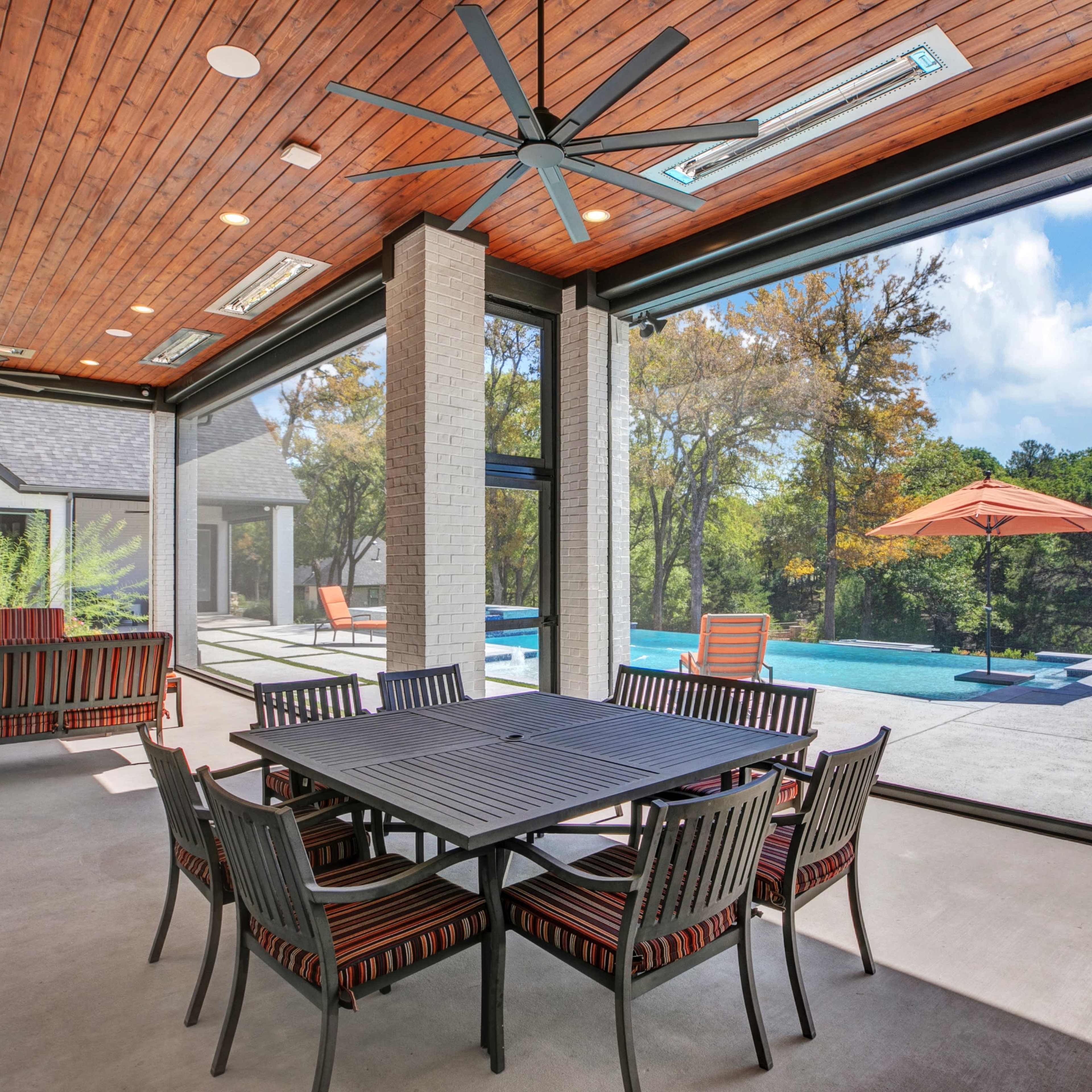 A covered outdoor patio features a dining table with chairs, ceiling fans, and large sliding glass doors that open to a pool area surrounded by trees.
