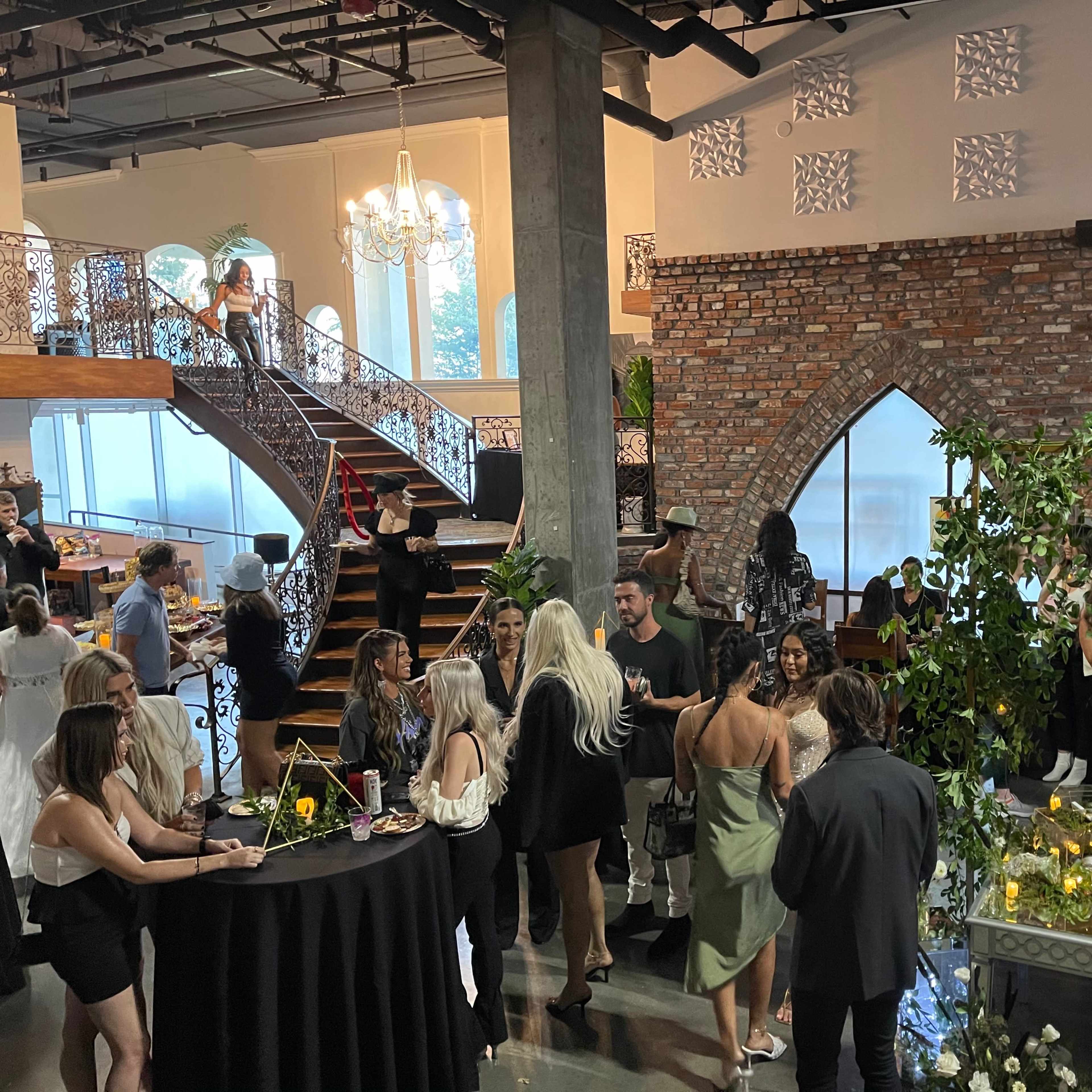 A group of people socializes at a stylish event held in a modern venue with exposed brick walls and elegant architecture.
