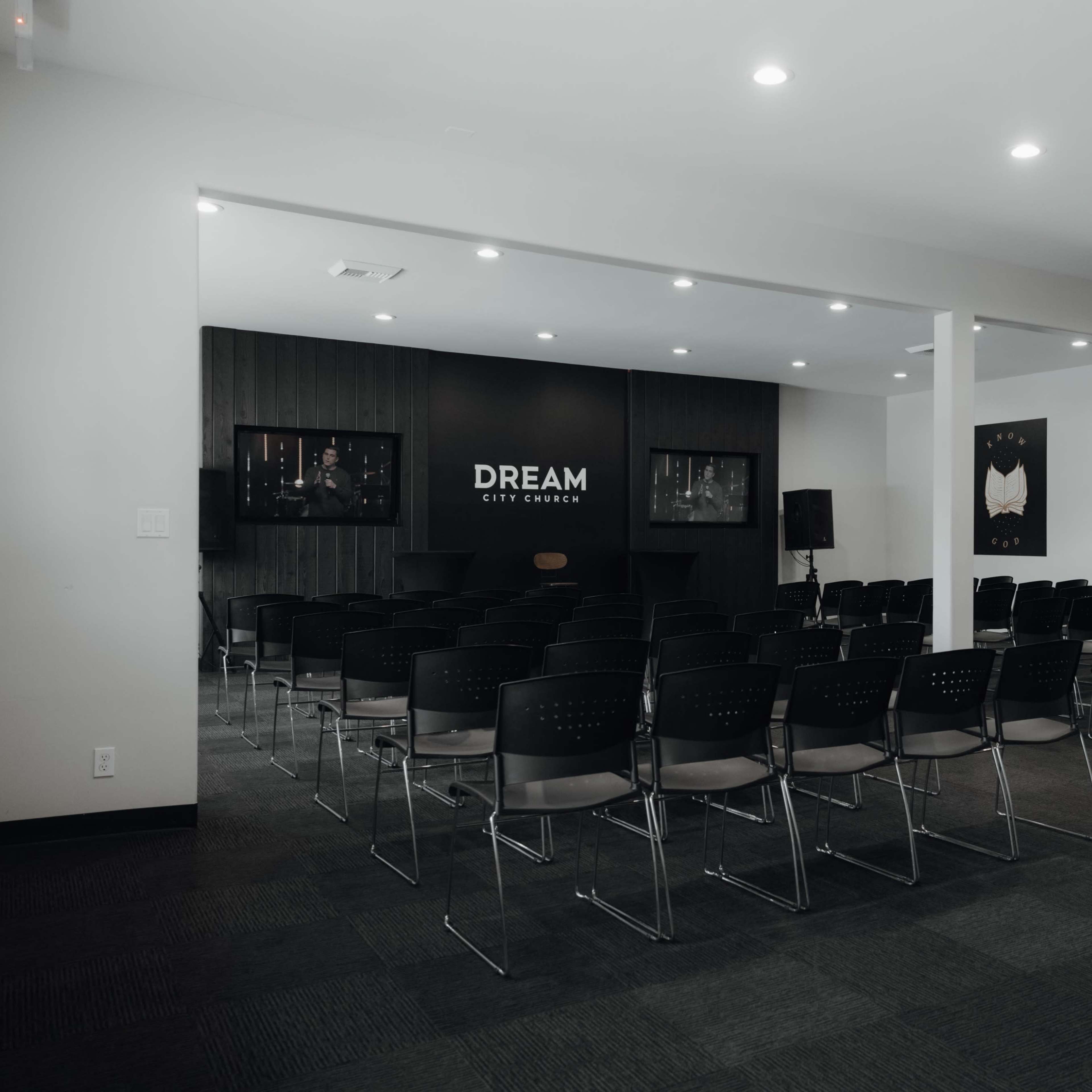 A conference room with rows of black chairs facing a stage that features a large sign reading "DREAM" and two artworks on the walls.