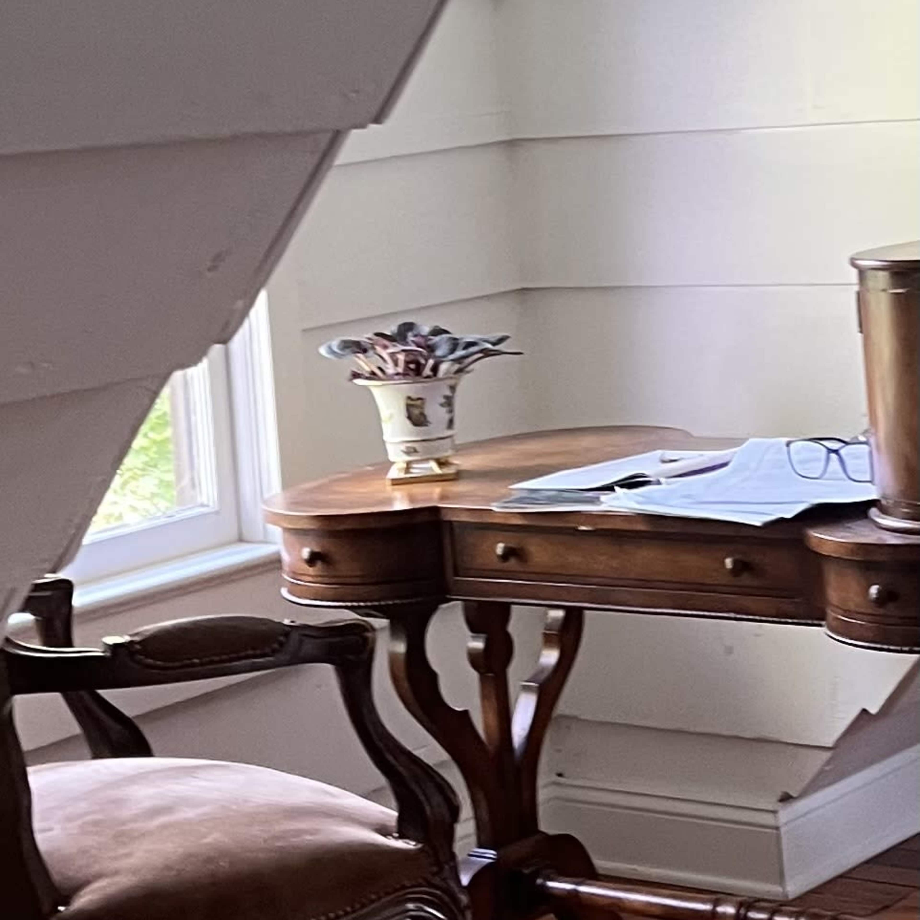 A wooden desk with papers and glasses rests beside a window, partially obscured by an angled wall.