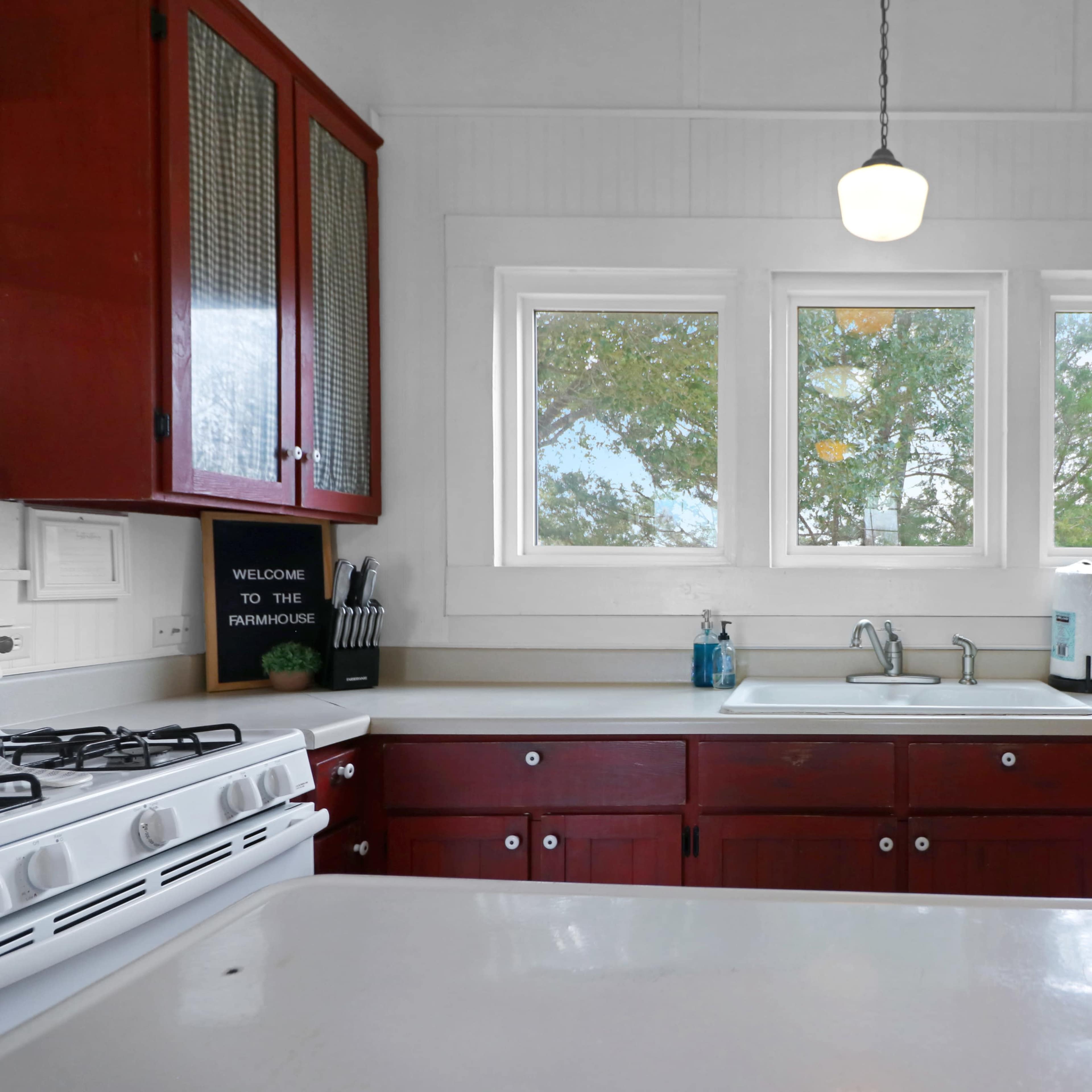 A kitchen features red cabinets and a white countertop, with a view through three windows overlooking greenery.