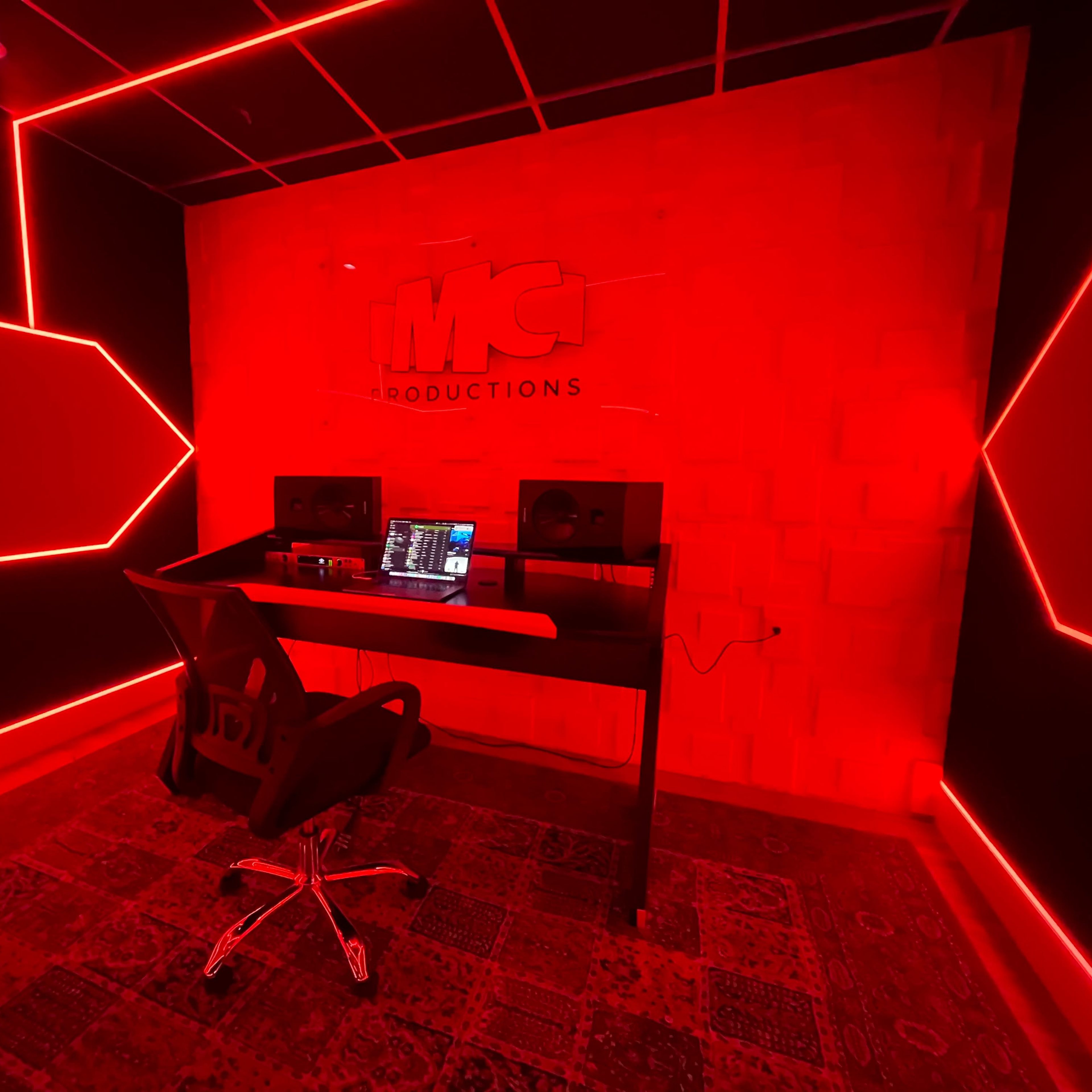 A music production studio features a black wall with red LED lighting, a desk with a laptop, and two studio monitors.