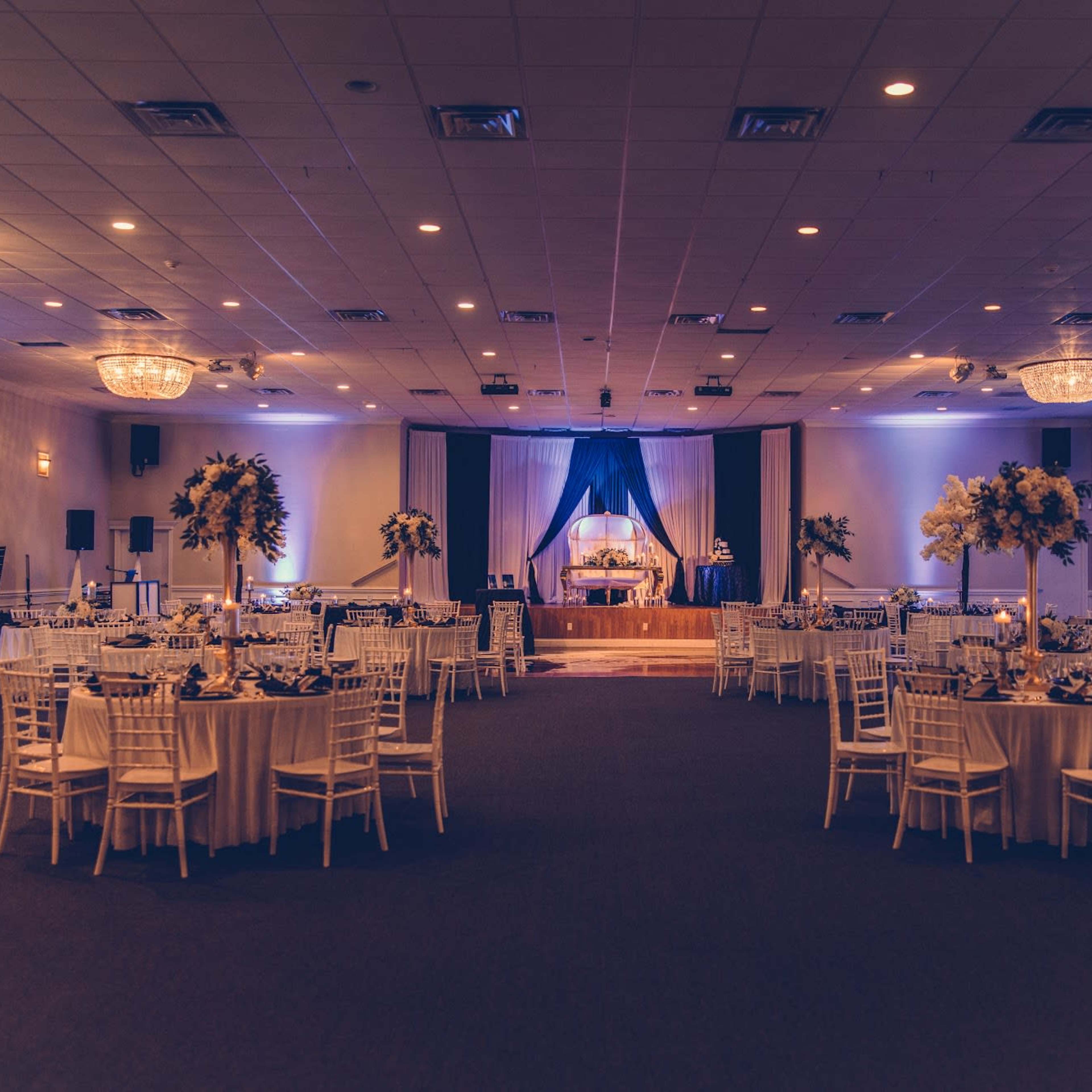 A spacious banquet hall is set up for an event, featuring round tables with white tablecloths, elegant centerpieces, and a staged area at the front adorned with blue drapery.