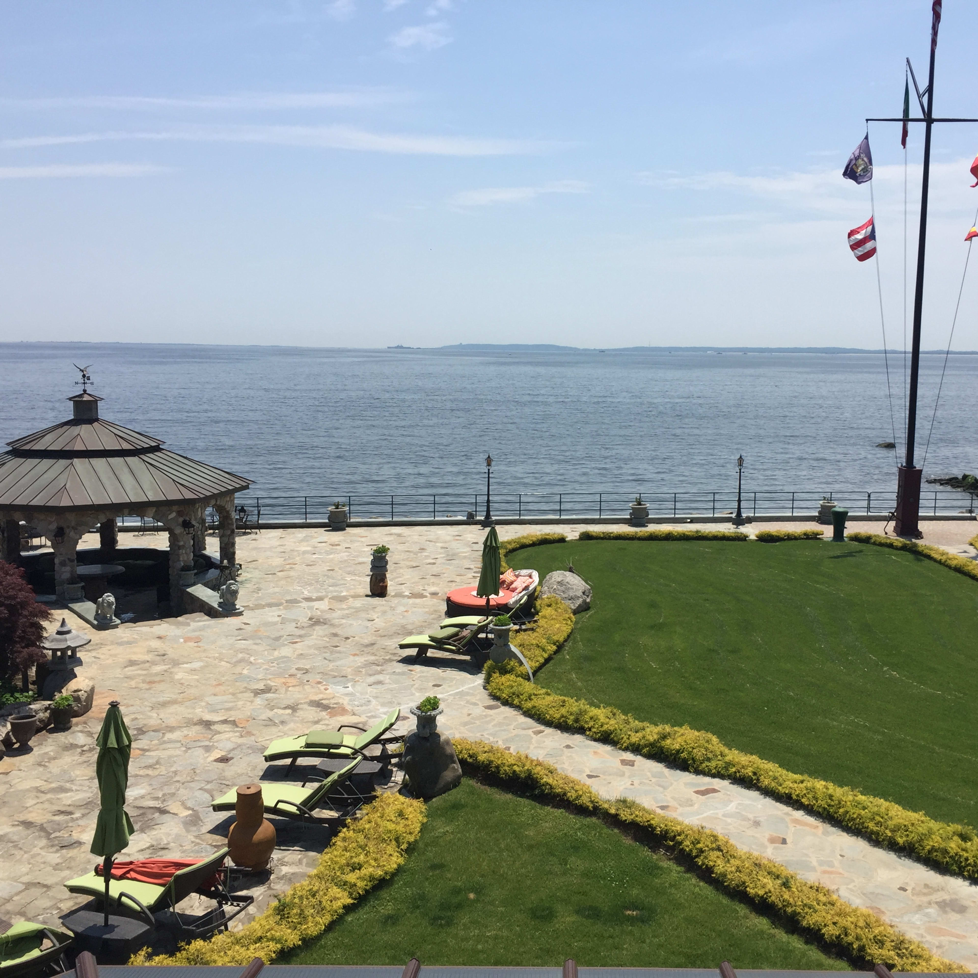 A stone-paved terrace overlooking a body of water, with lounge chairs and flags nearby.