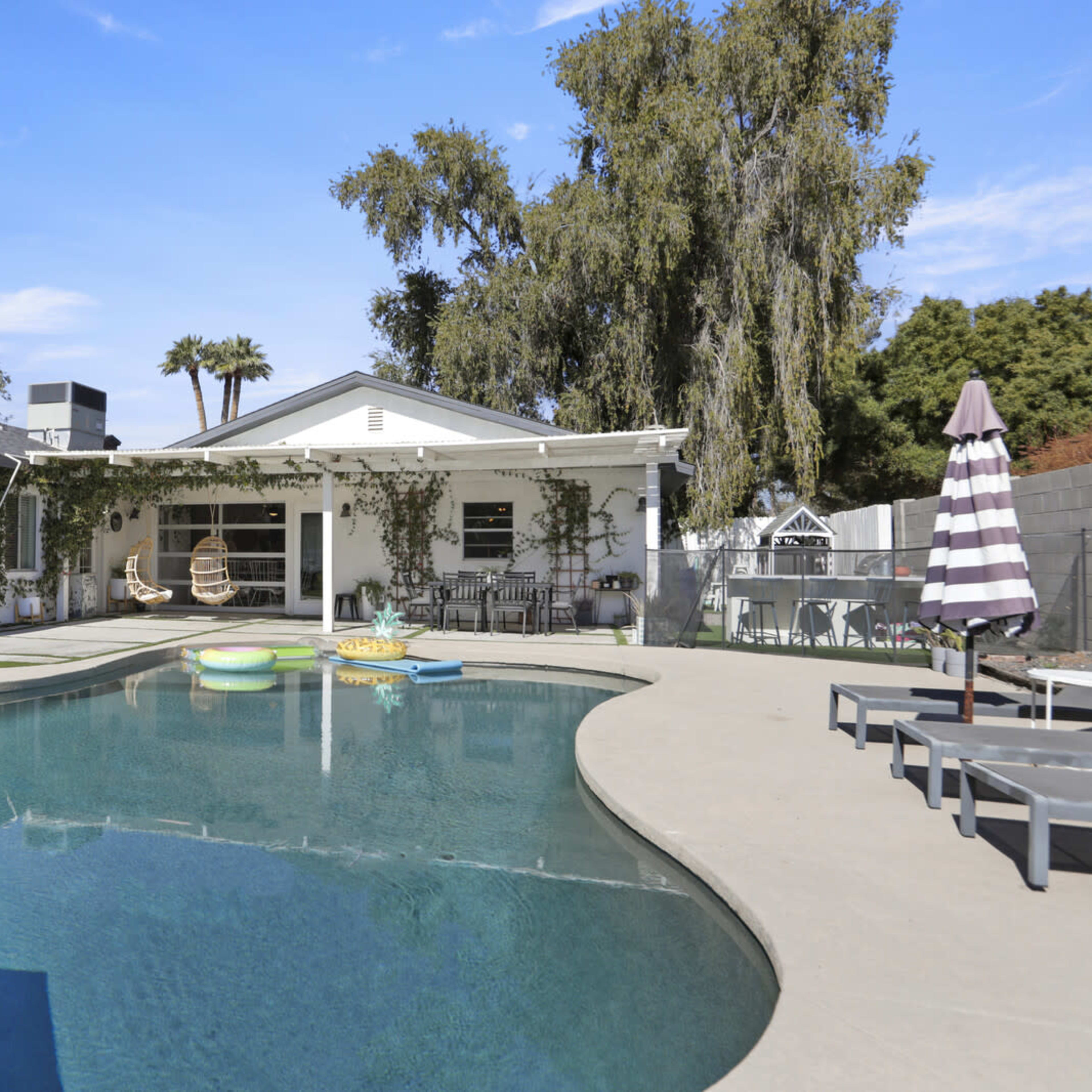 A backyard features a pool with floating objects, surrounded by lounging chairs and a covered patio with outdoor seating.