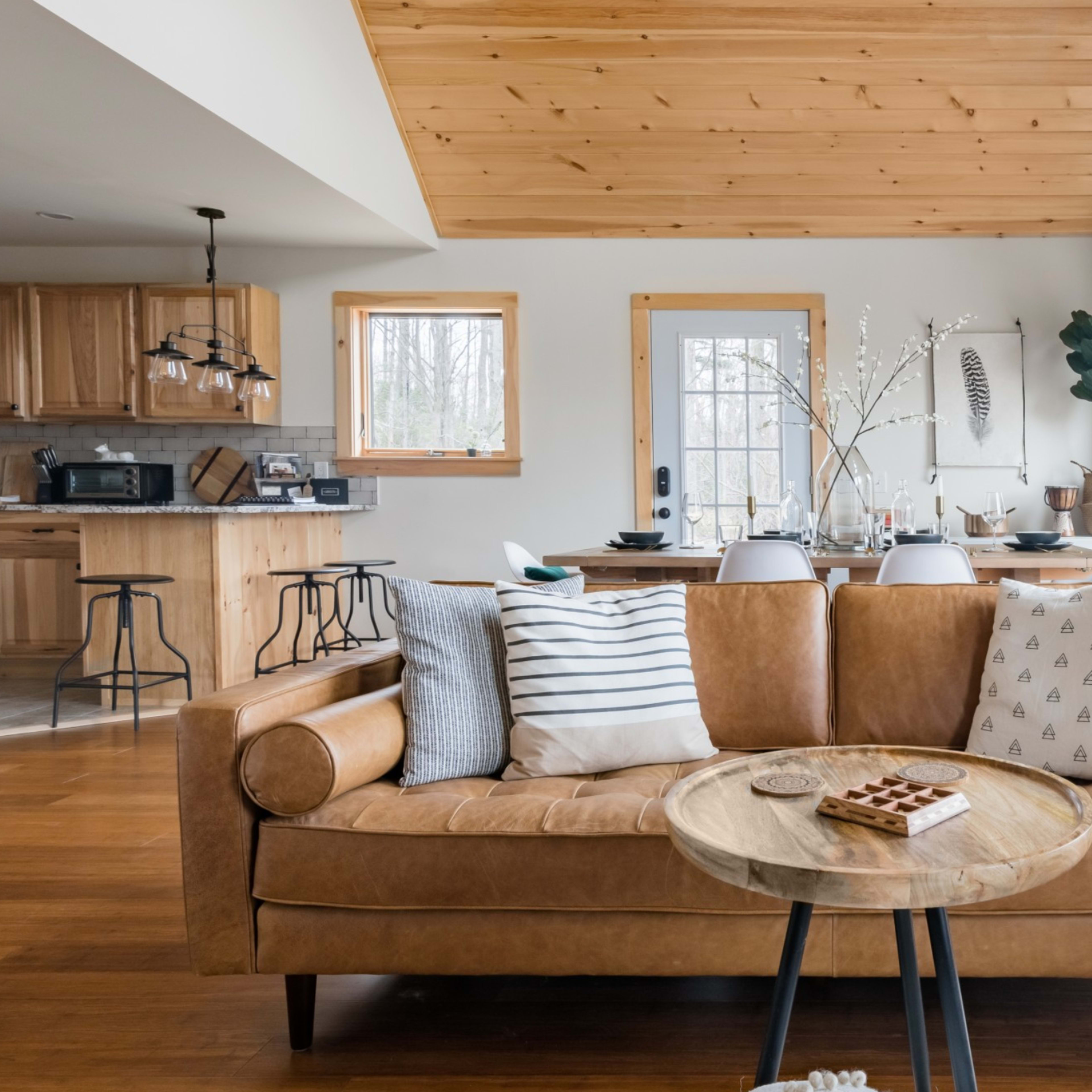 A cozy living room with a leather sofa, a wooden coffee table, and large windows leading to a deck, with a kitchen visible in the background.