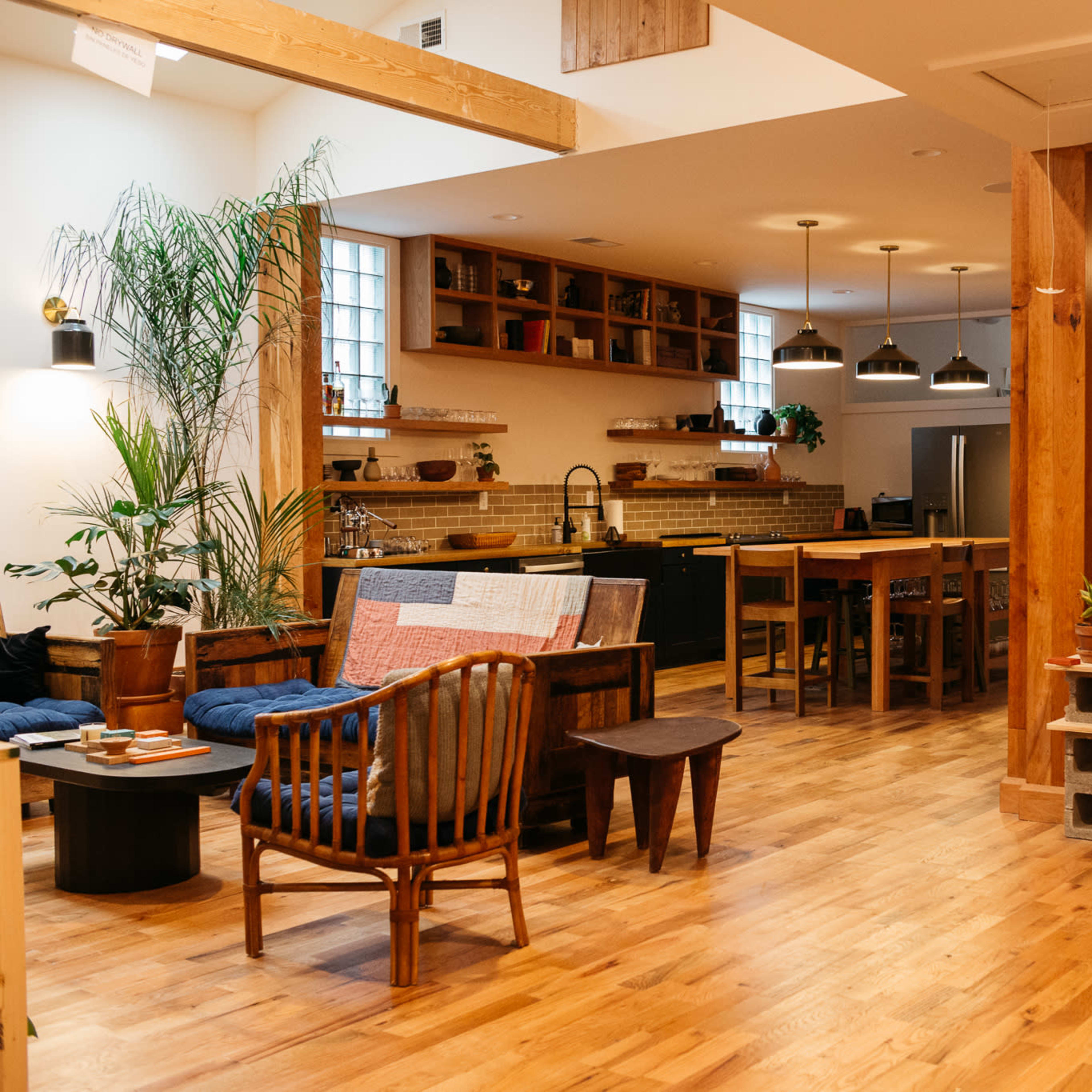 A spacious, open-plan living area featuring wooden floors, a kitchen with a dining table, and various plants arranged throughout.