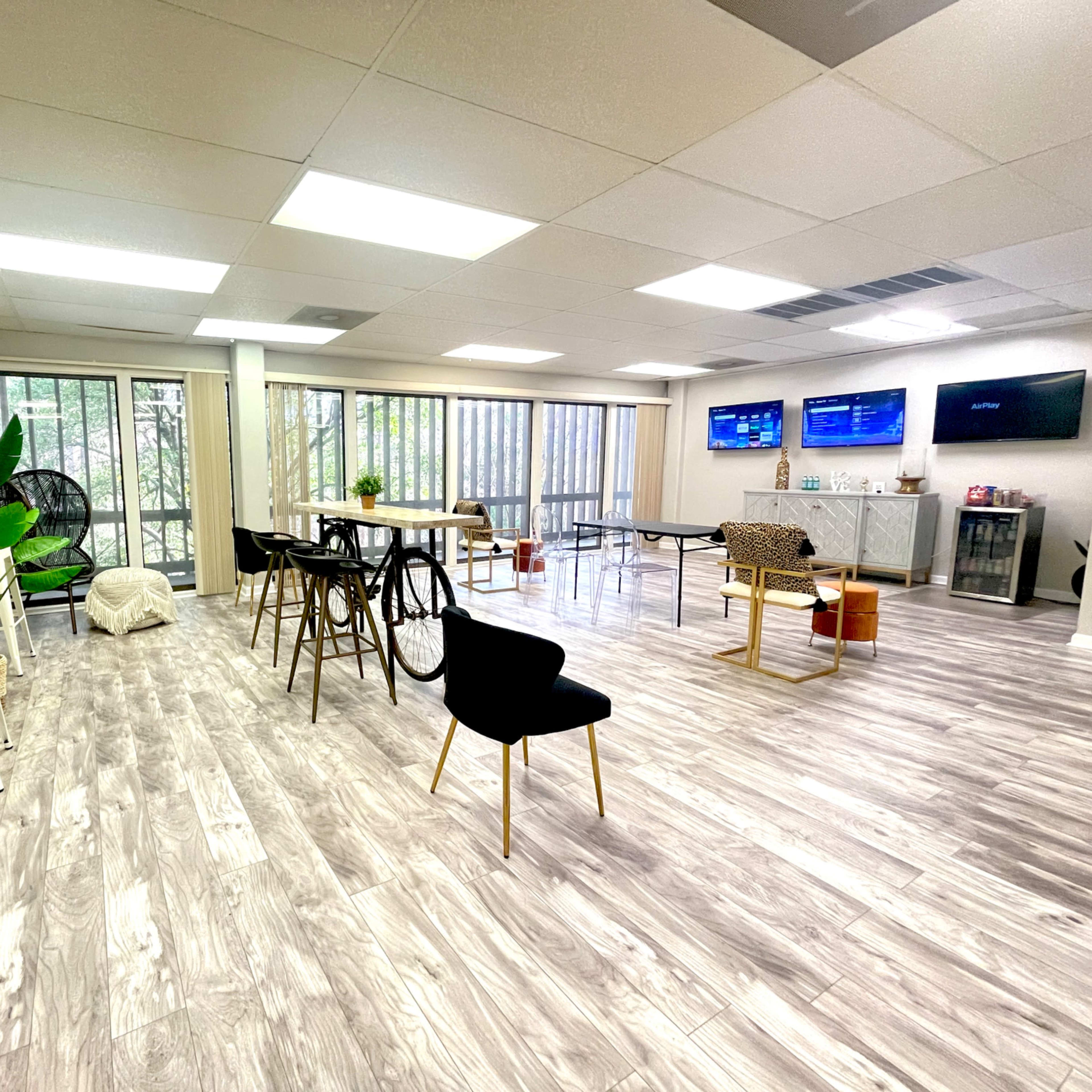 A spacious, well-lit room with multiple tables, chairs, a large window with greenery outside, and several wall-mounted displays.