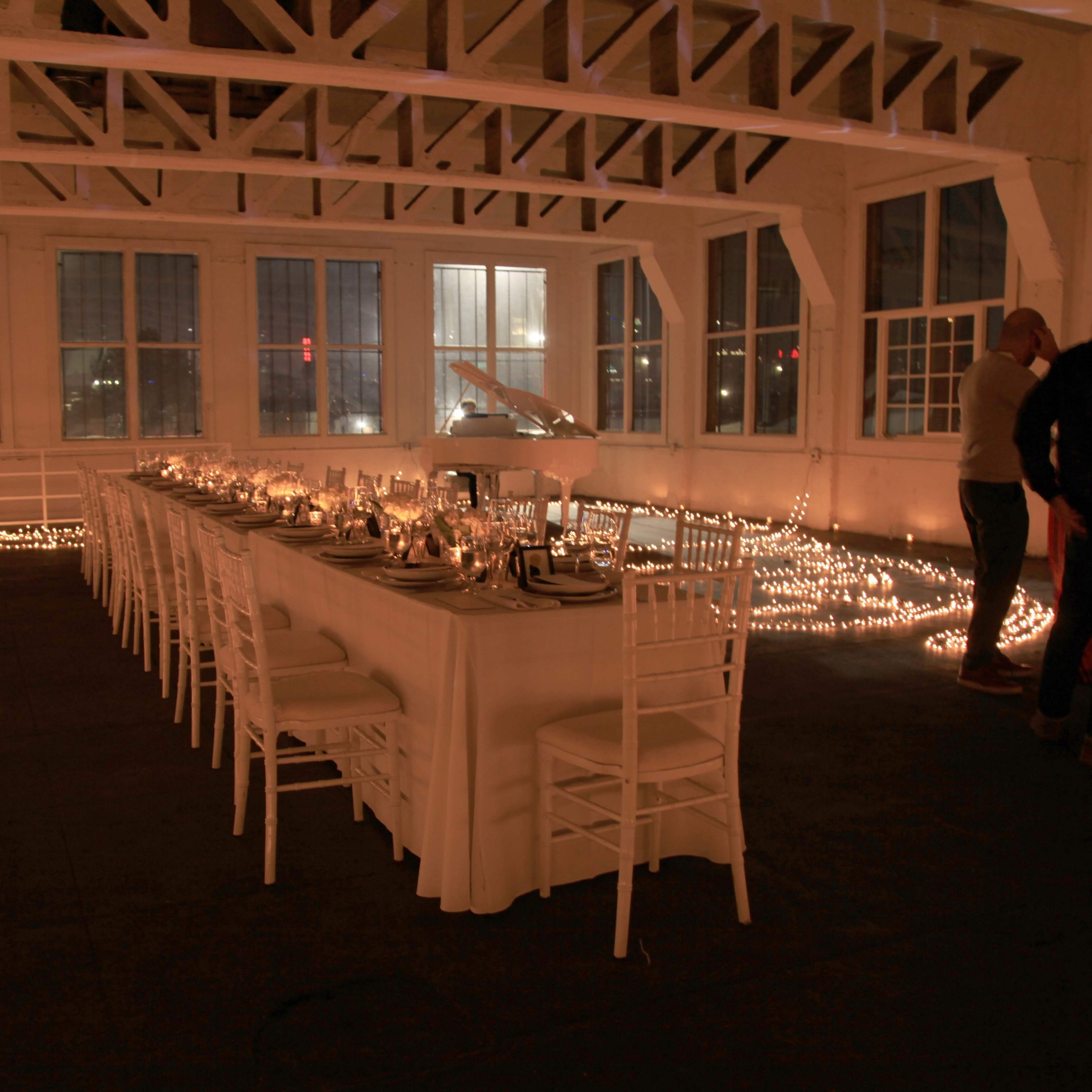 A long dining table is set in a well-lit, airy room adorned with string lights along the floor, while guests mingle nearby.