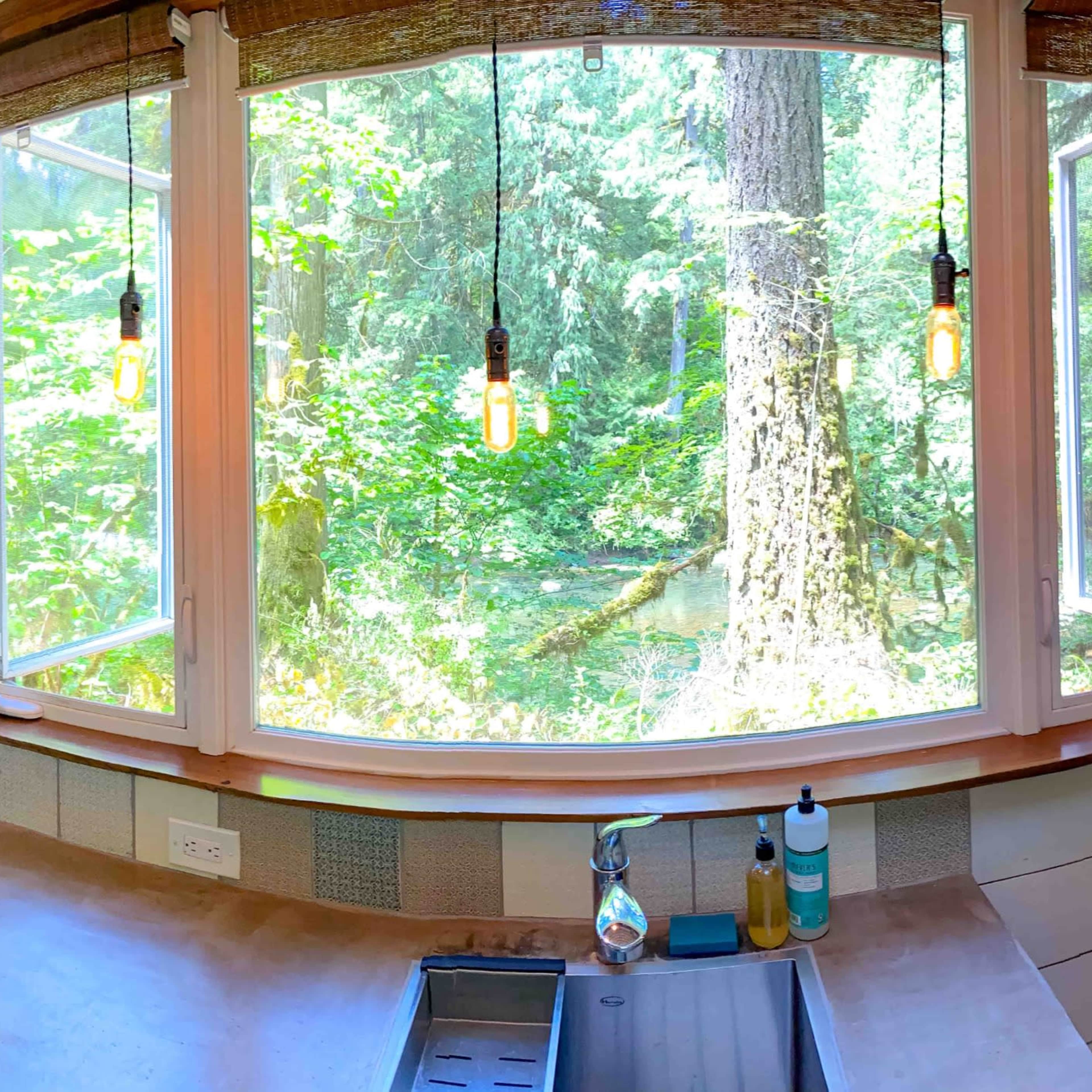 A cozy kitchen area with large windows overlooking a forest, featuring a stove, sink, and a seating nook with cushions.