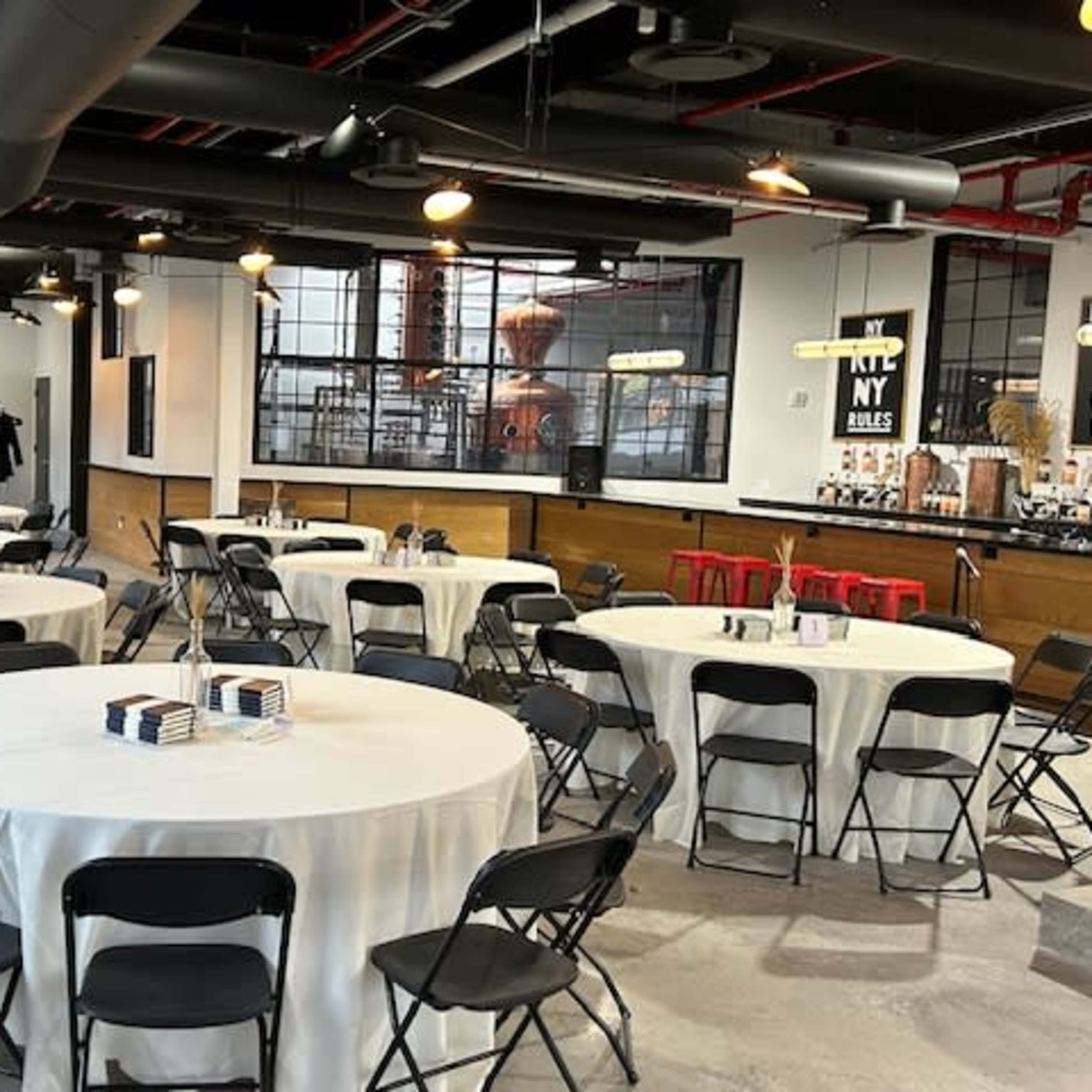A neatly arranged event space with several round tables covered with white tablecloths and black chairs, featuring a bar area in the background.
