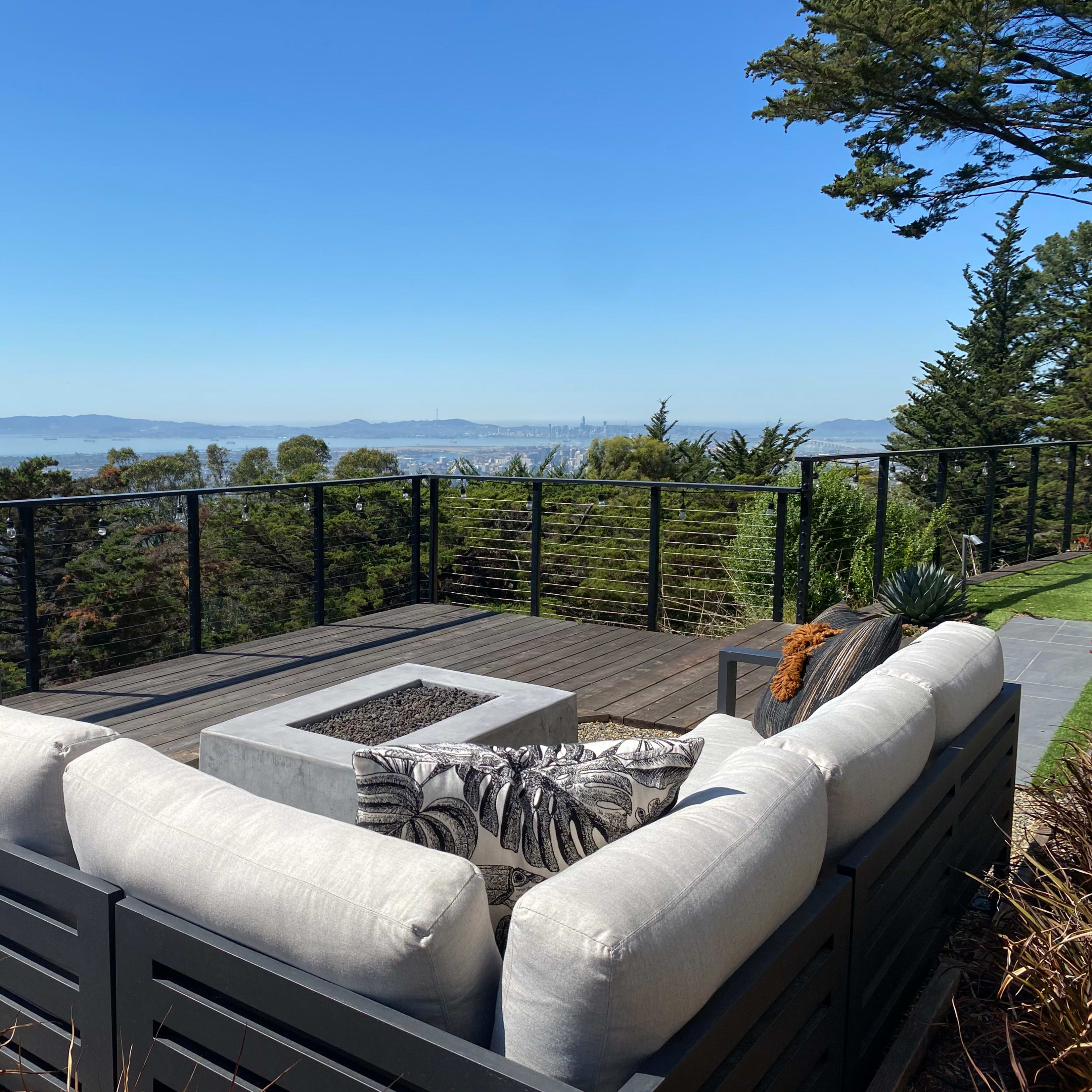 A modern outdoor lounge area with a fire pit overlooks a city and bay through a railing of trees.