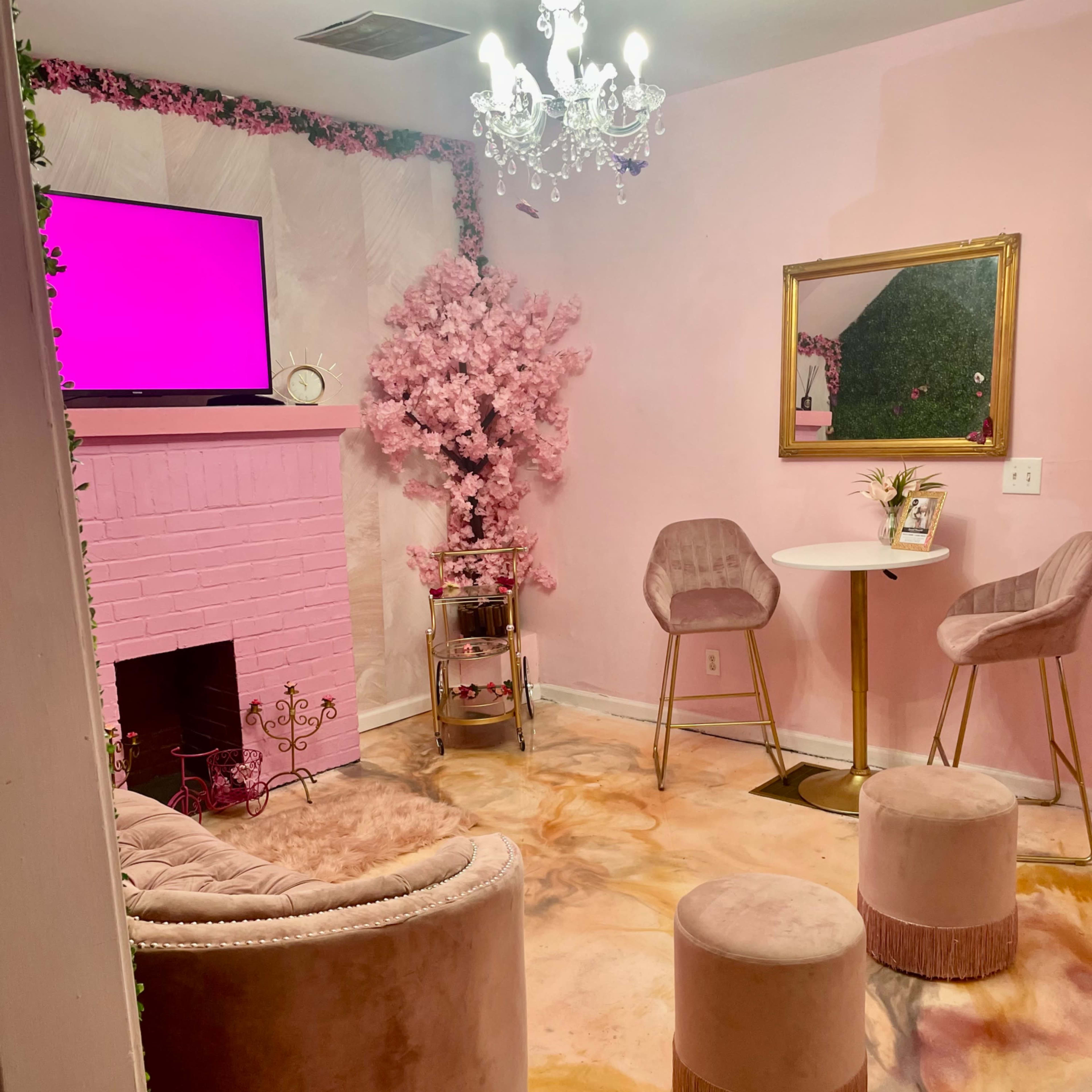 A cozy, pink-themed room featuring a TV on the wall, a chandelier, a faux cherry blossom tree, and seating arrangements of plush chairs and stools on a colorful floor.