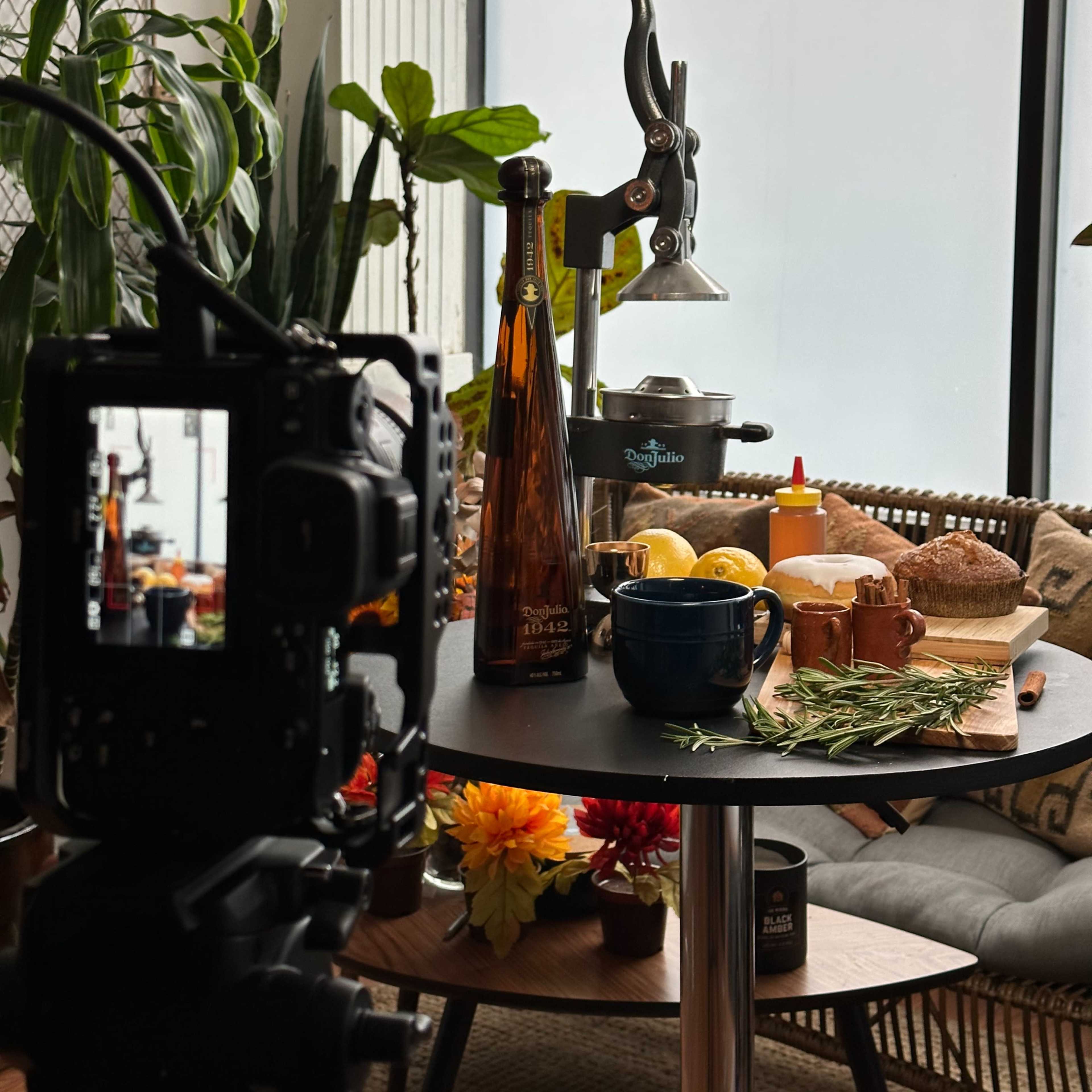 A tabletop arrangement with a bottle, a citrus juicer, sliced lemons, a cup, and herbs, with a camera focused on the scene.