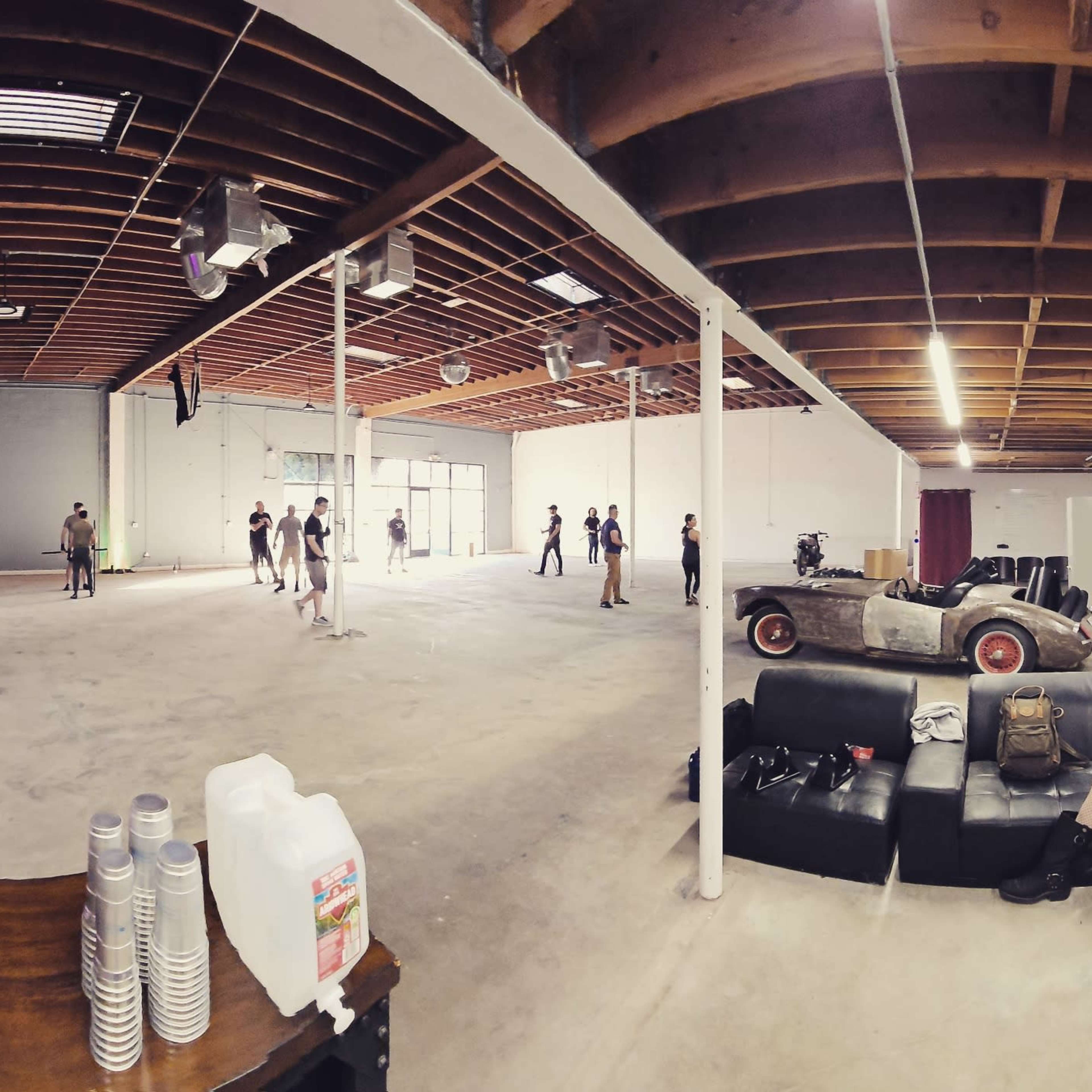 A spacious, unfinished indoor environment with a group of people engaging in activities, a ladder positioned to the side, and various furniture and equipment scattered around.