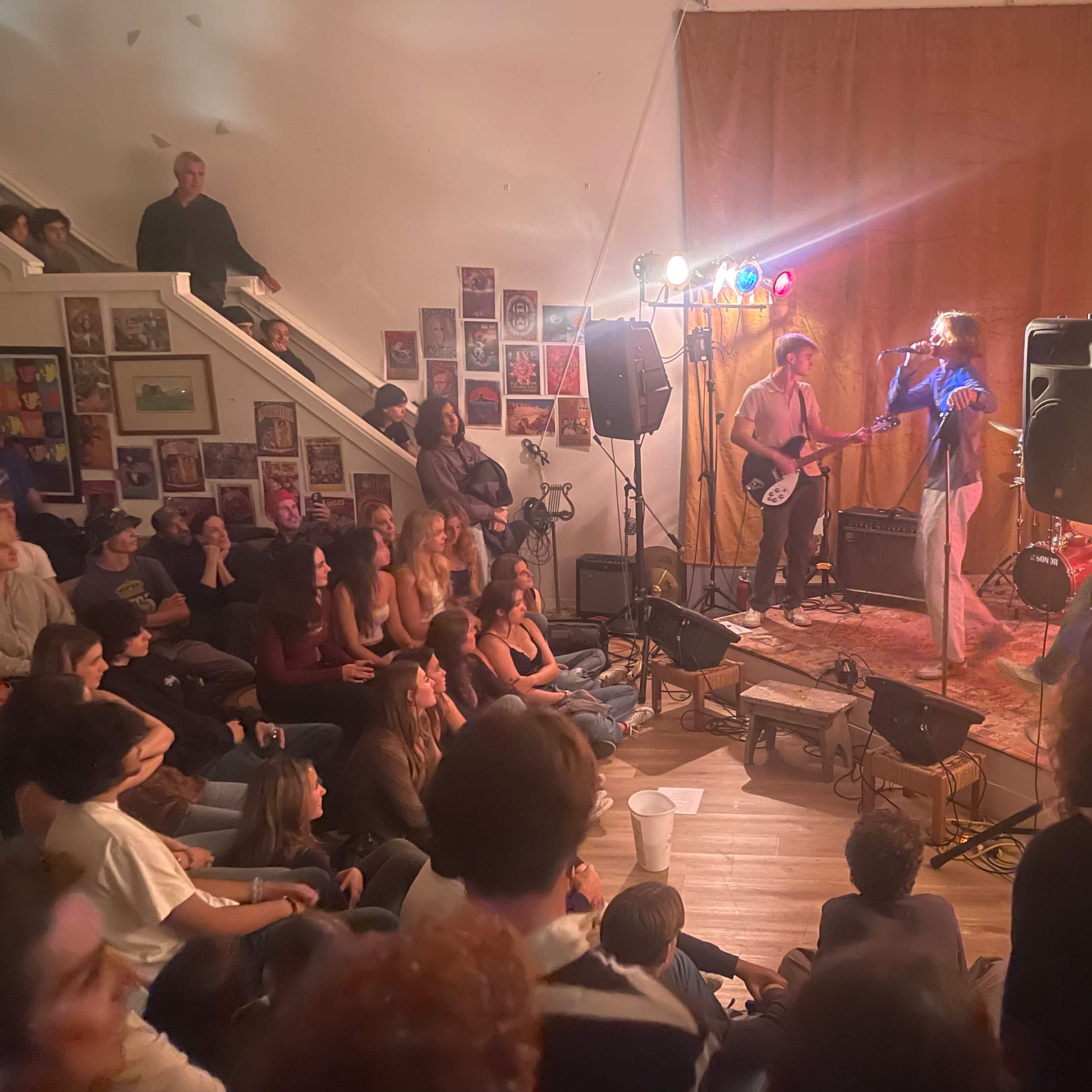 A live music performance is taking place in a crowded room with an audience sitting on the floor and standing around a small stage.