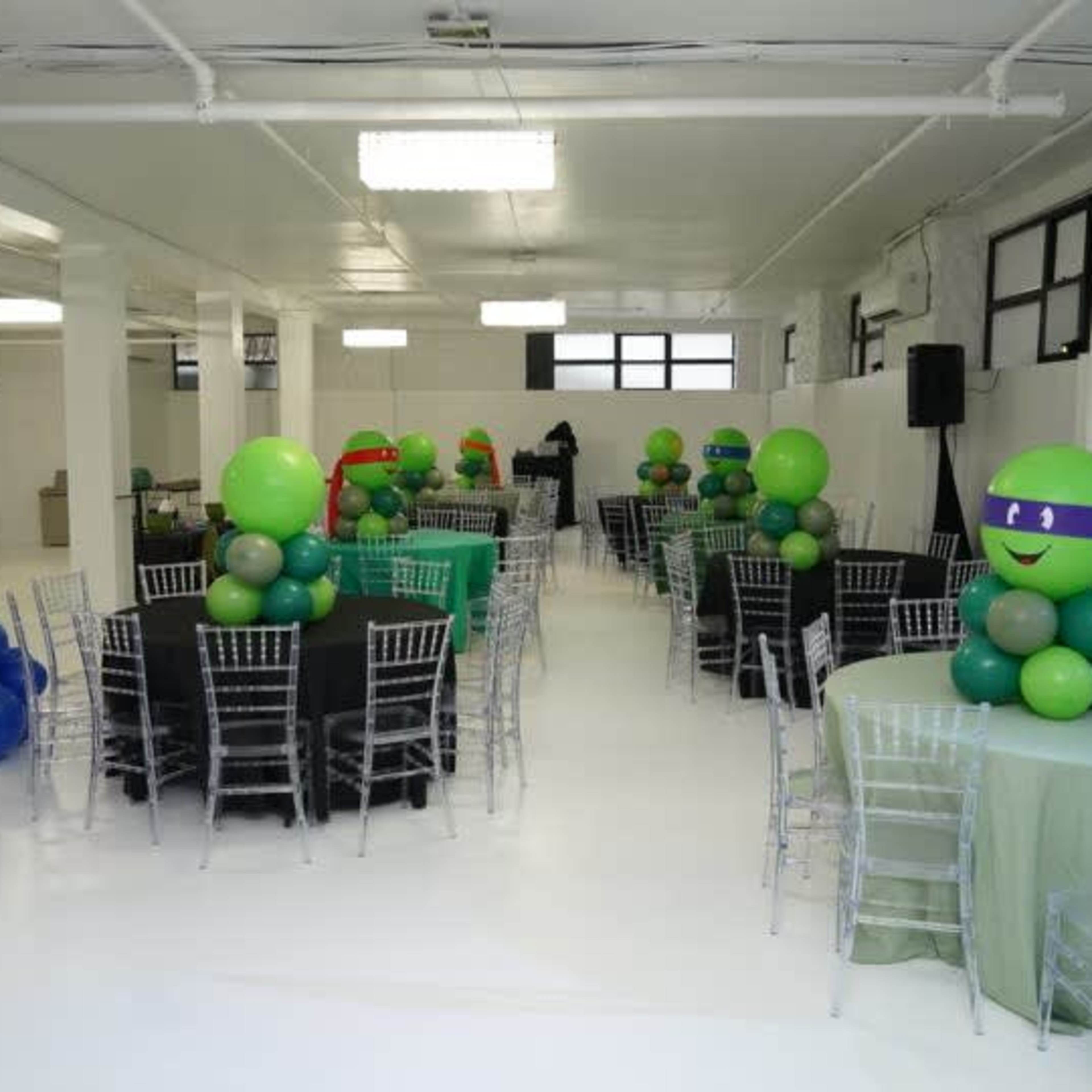 A bright event space decorated with tables covered in black and green tablecloths, featuring balloon centerpieces of varying colors, including green and purple.