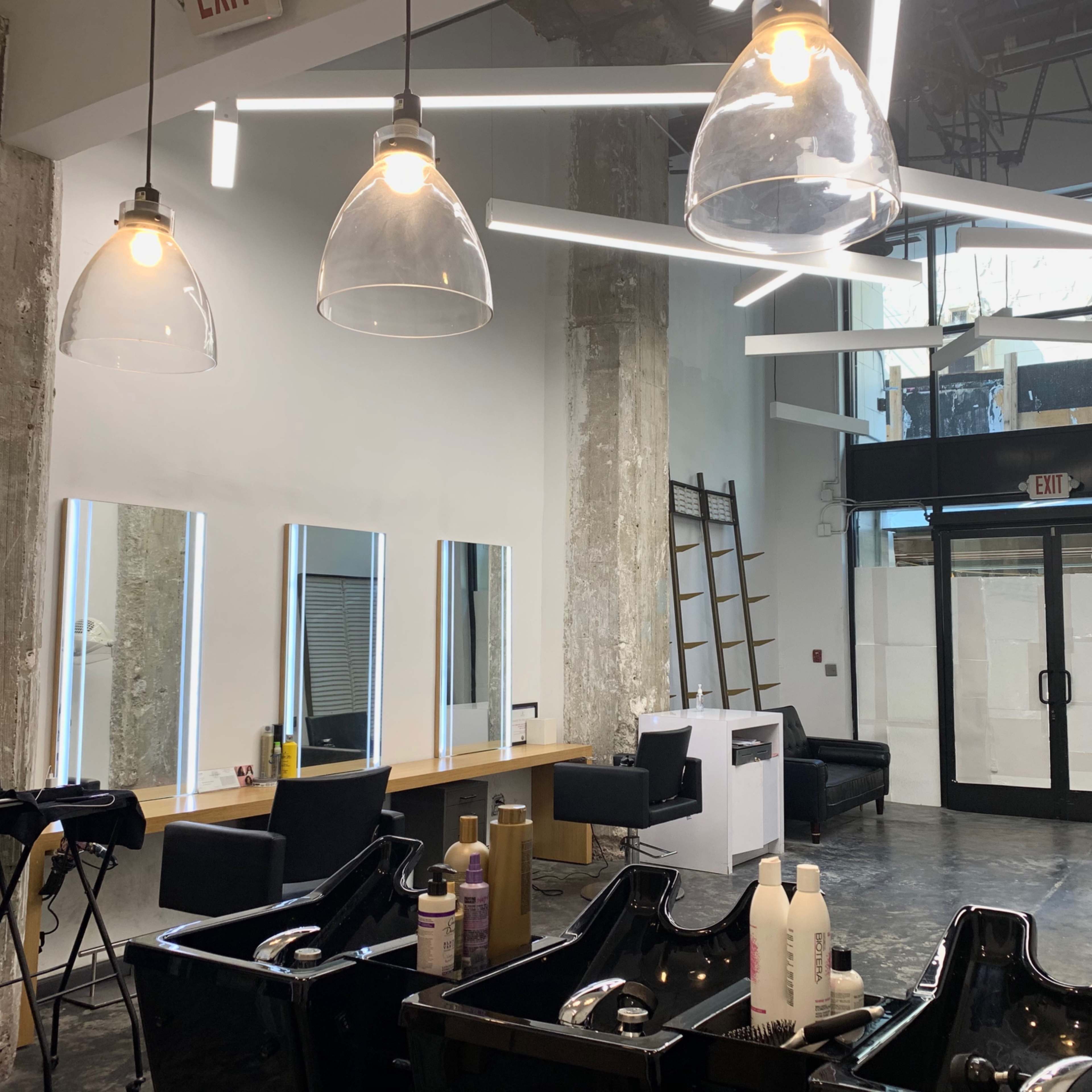 The interior of a modern salon features black shampoo bowls, illuminated mirrors, and a sleek entrance with large glass doors.