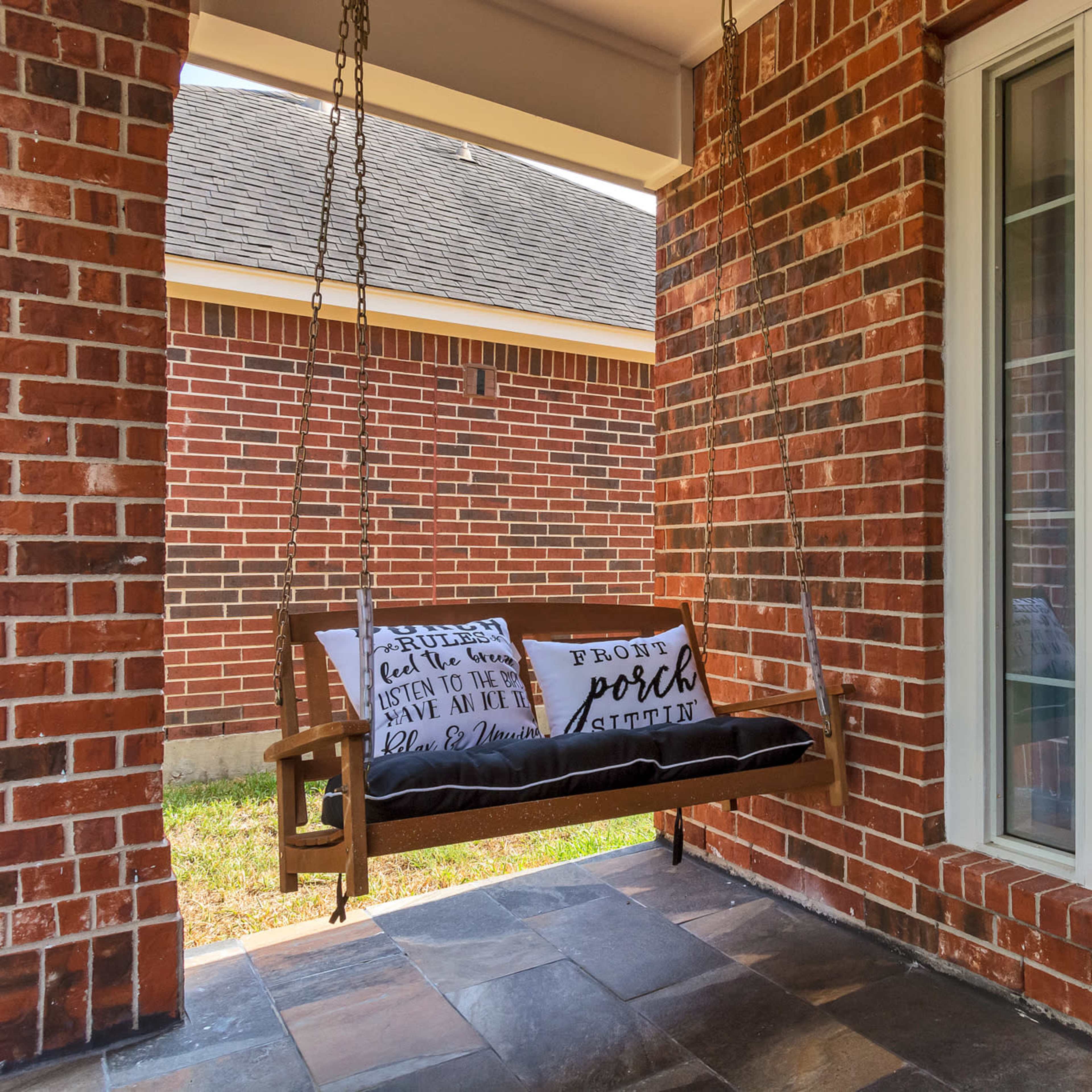 A wooden swing with decorative pillows hangs on a front porch made of brick and stone.